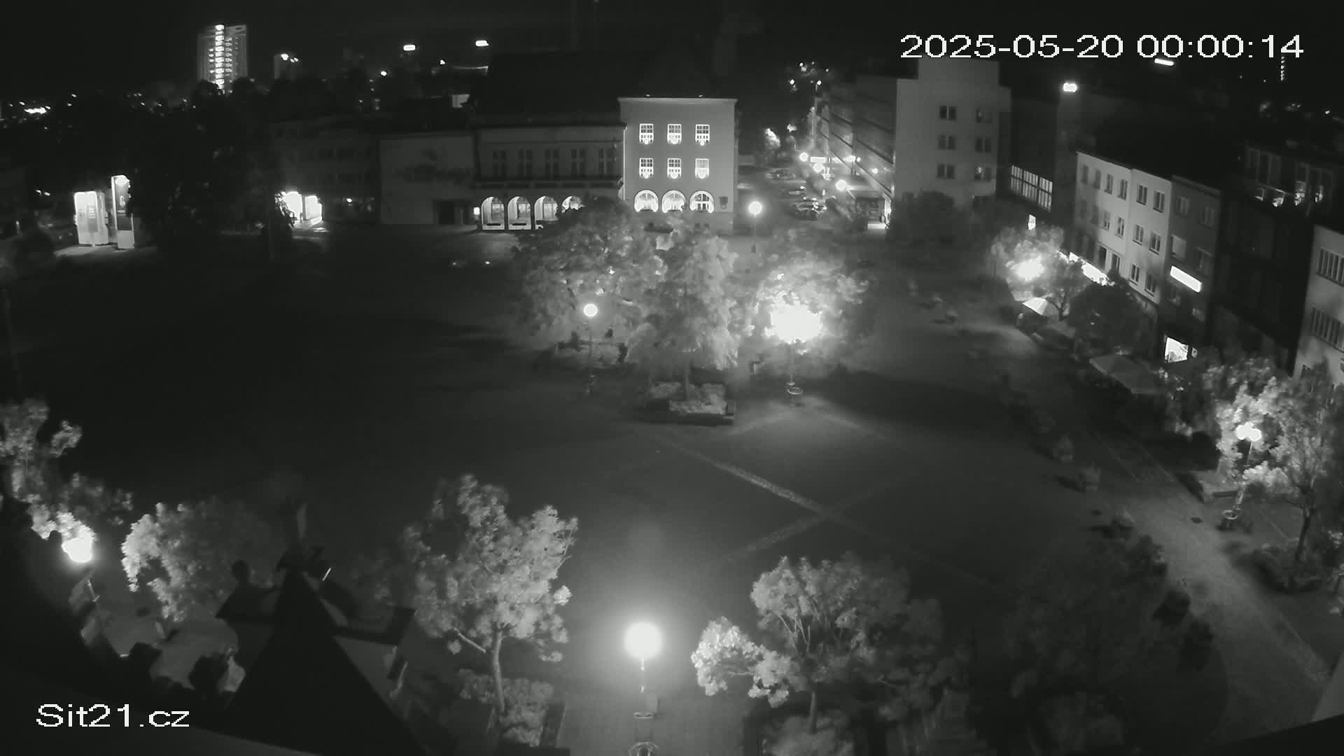 A nighttime, overhead view of a town square with buildings surrounding a sparsely populated area illuminated by streetlights.