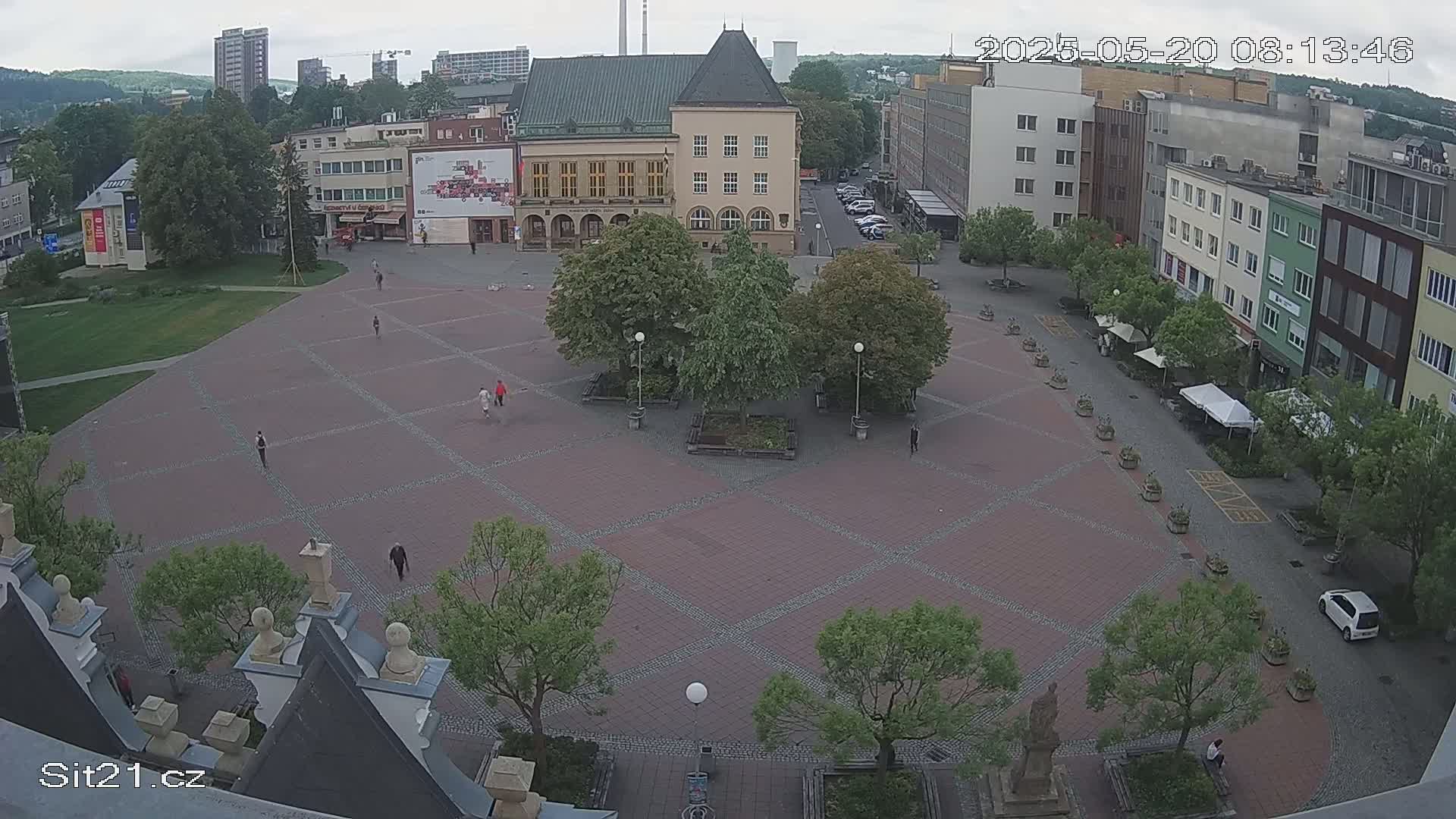 A mostly empty town square with a brick pavement, several trees, and buildings surrounding it under an overcast sky.