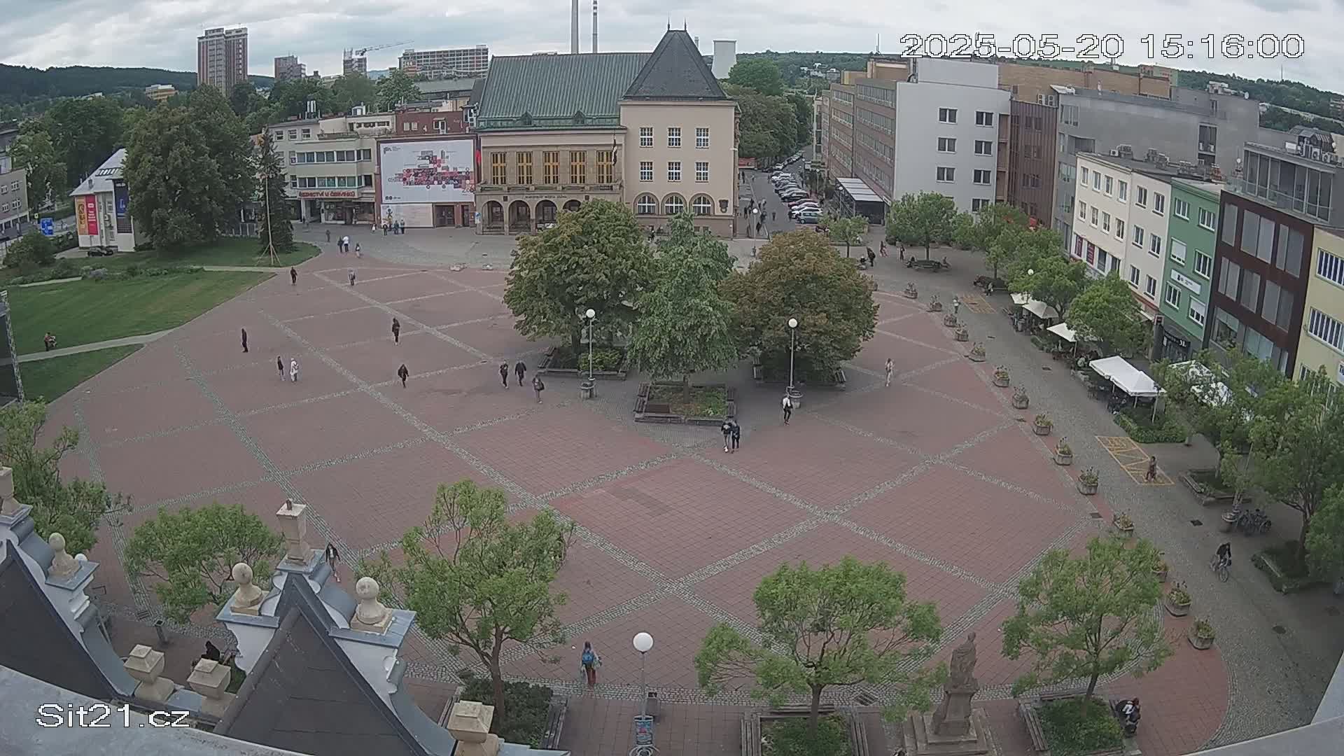 A mostly empty brick town square with several trees and buildings surrounding it, under a cloudy sky.