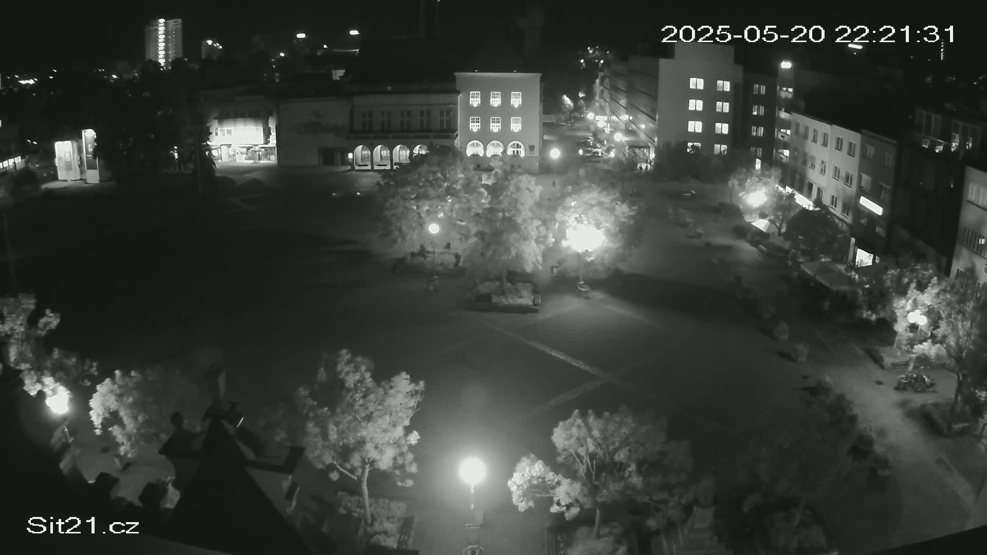 A nighttime, dimly lit, empty town square is surrounded by buildings and trees, illuminated by several street lamps.
