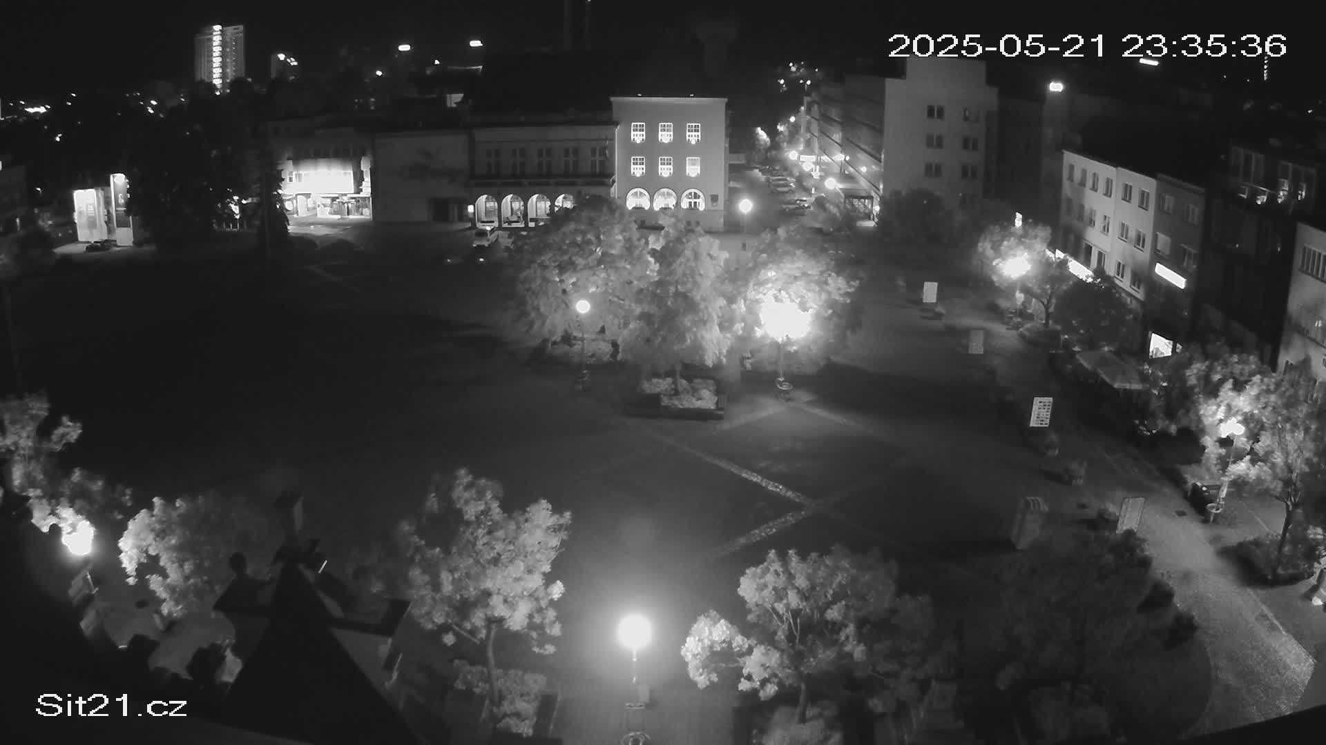 A nighttime, high-angle view of a town square surrounded by buildings and trees, illuminated by streetlights.
