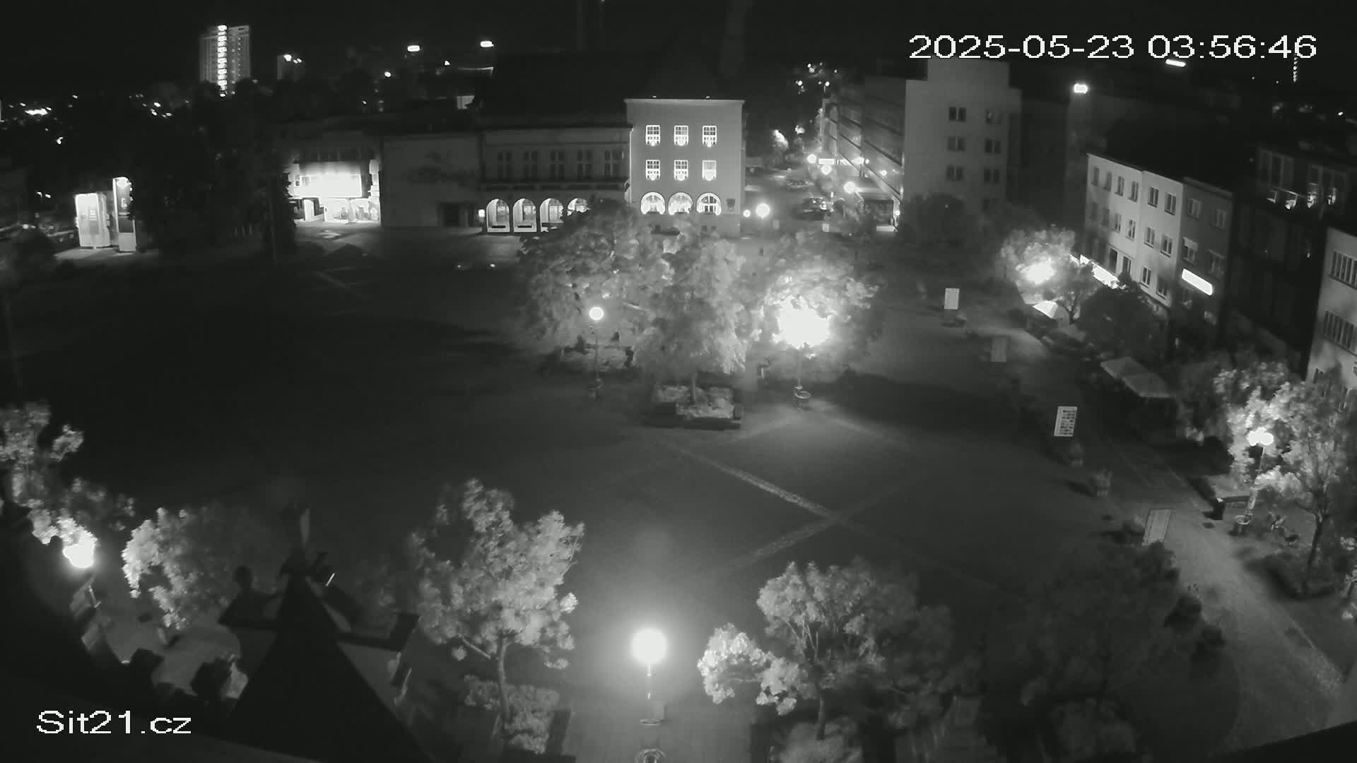 A nighttime, high-angle view of a town square with several buildings, trees, and streetlights illuminating the scene.