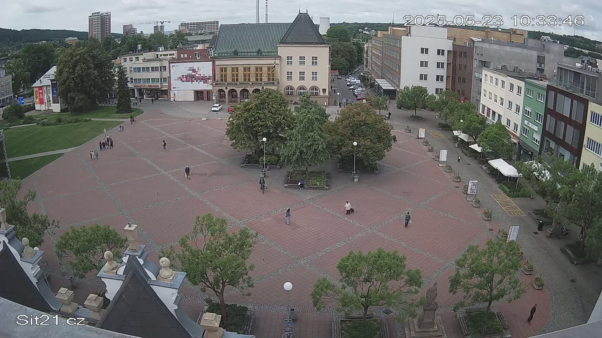 A brick town square with several people walking around, surrounded by buildings and green trees under an overcast sky.
