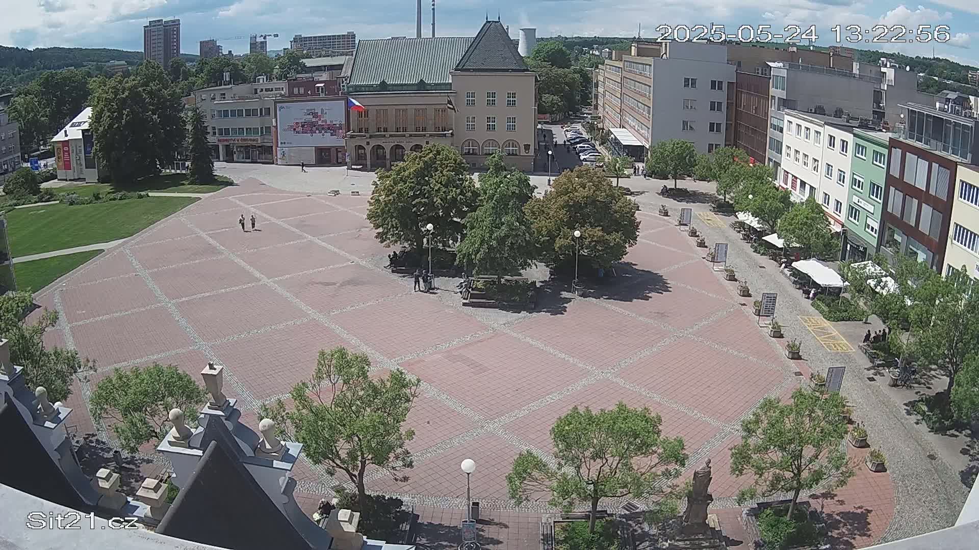 A brick town square with several trees and a few people is seen from an elevated viewpoint on a partly sunny day.