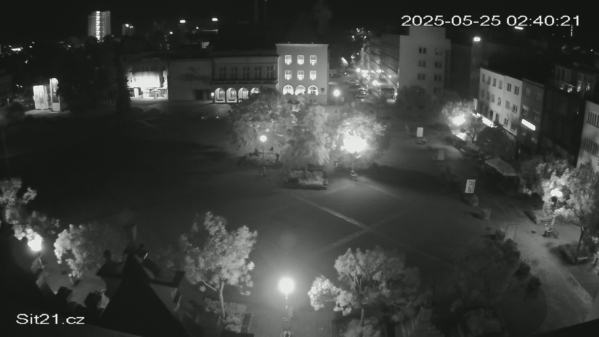 A nighttime, high-angle view of a town square with buildings surrounding it, illuminated by streetlights and featuring trees and some sparsely distributed people.