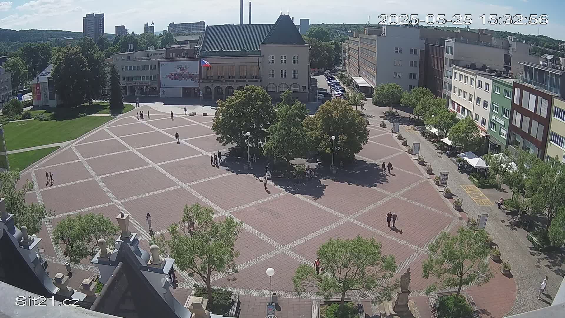 A large, paved town square with several people walking around, surrounded by buildings and trees under a sunny sky.