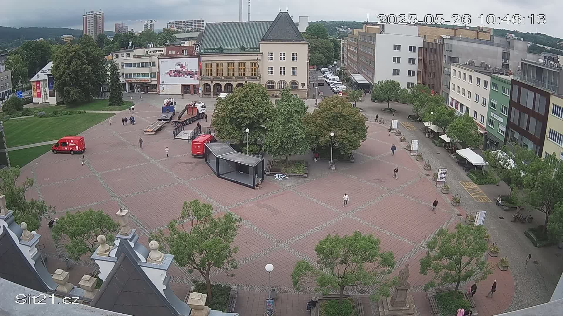 A brick town square with several people walking around, some trees, and a few red trucks and vans, under an overcast sky.