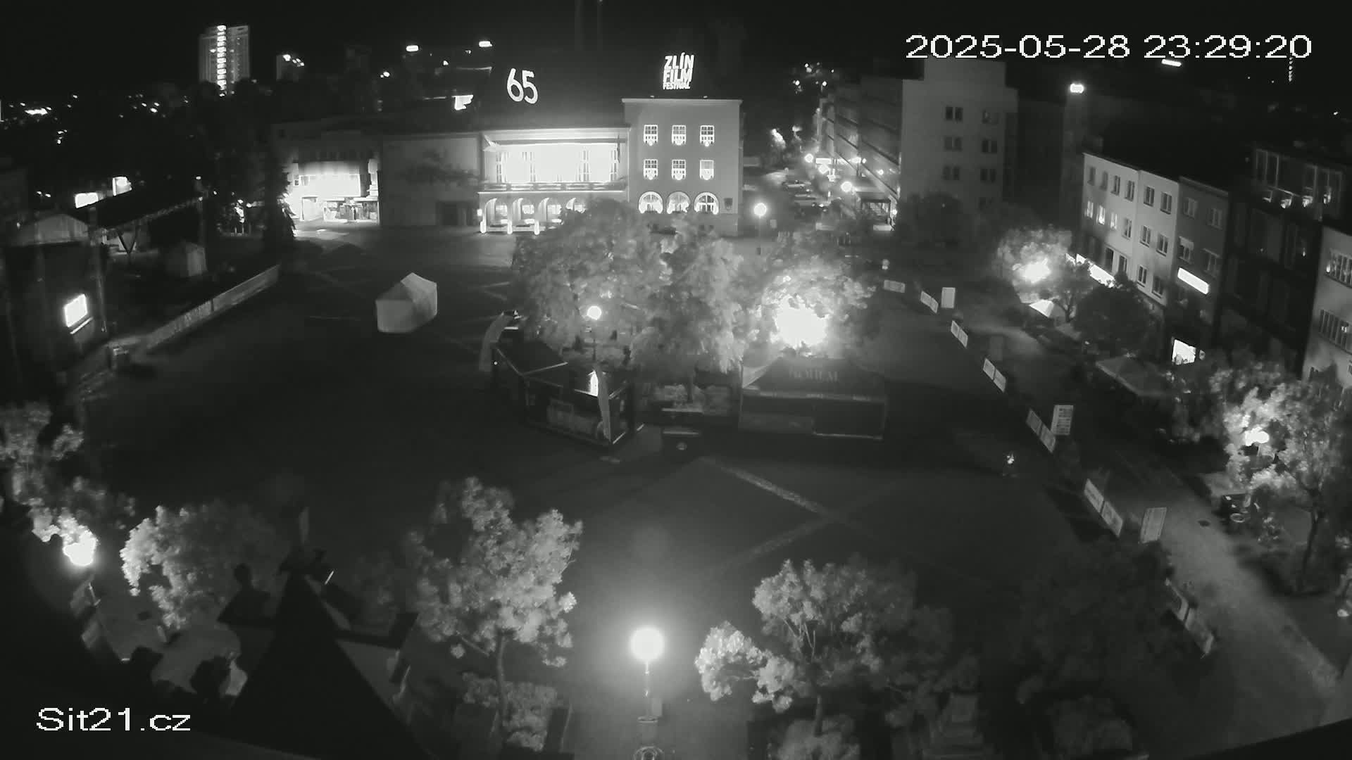 A nighttime, high-angle, black-and-white view of a city square with buildings, trees, and some structures that appear to be temporary vendor stalls or booths.