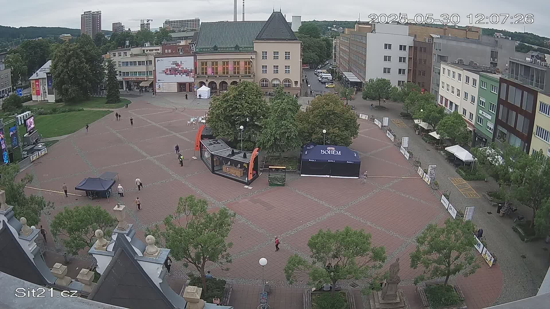 An overcast day reveals a large, brick-paved town square with several small tents, kiosks, and a few people sparsely scattered throughout, surrounded by multi-story buildings.