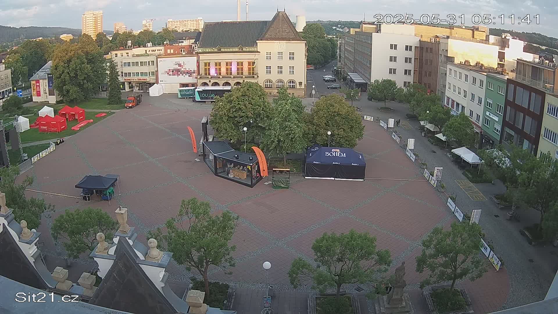 An overcast outdoor town square with several tents, buildings, and trees, under a partly cloudy sky.
