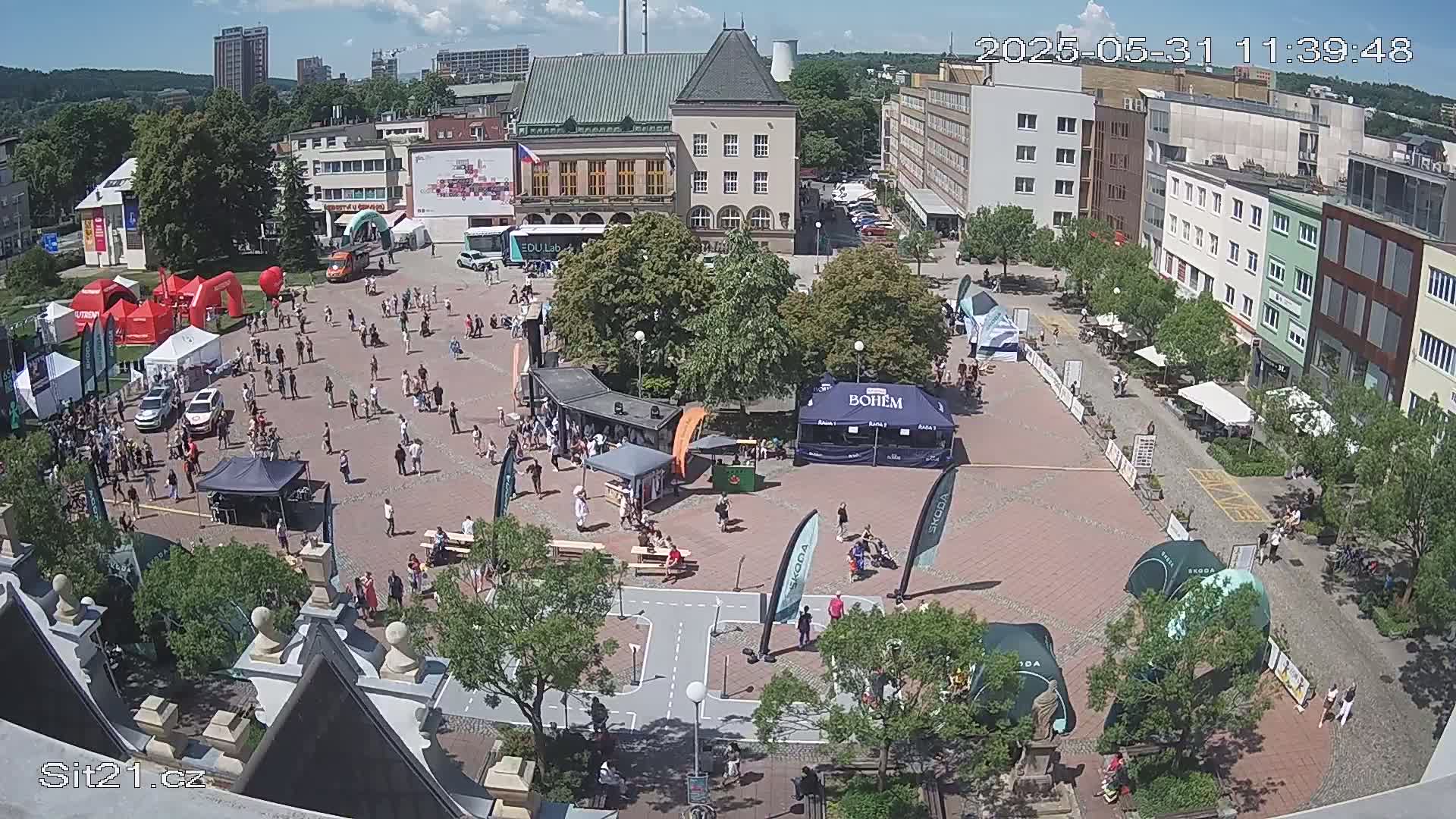 A town square with many people and numerous tents and booths under a mostly sunny sky.