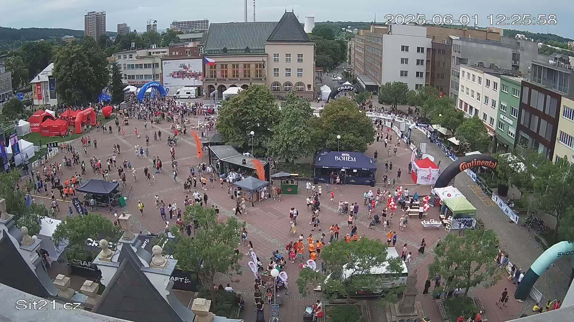A large crowd of people gathers in a town square on a partly cloudy day, with various booths and inflatable structures set up, suggesting a festival or event.