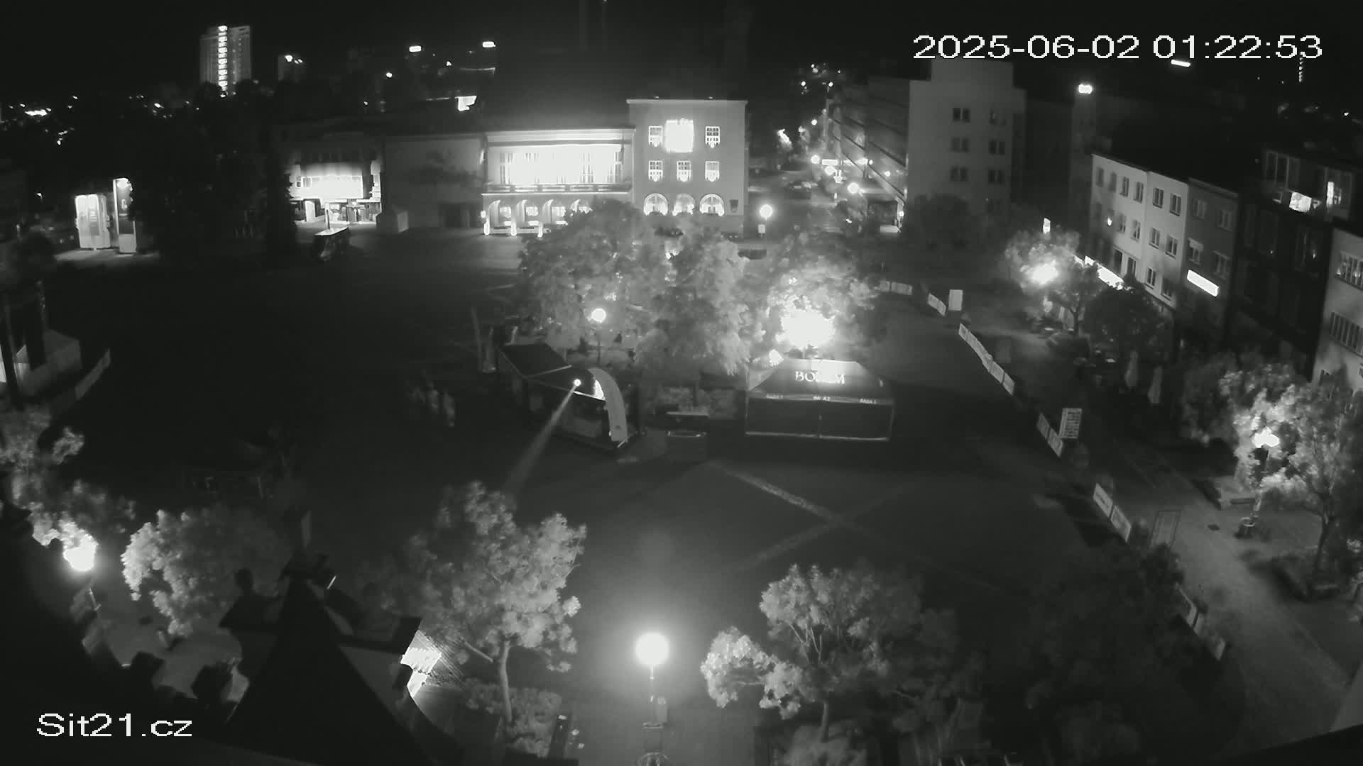 A nighttime, high-angle view of a town square with buildings surrounding it, lit by streetlights and featuring some sparsely populated areas under a clear sky.