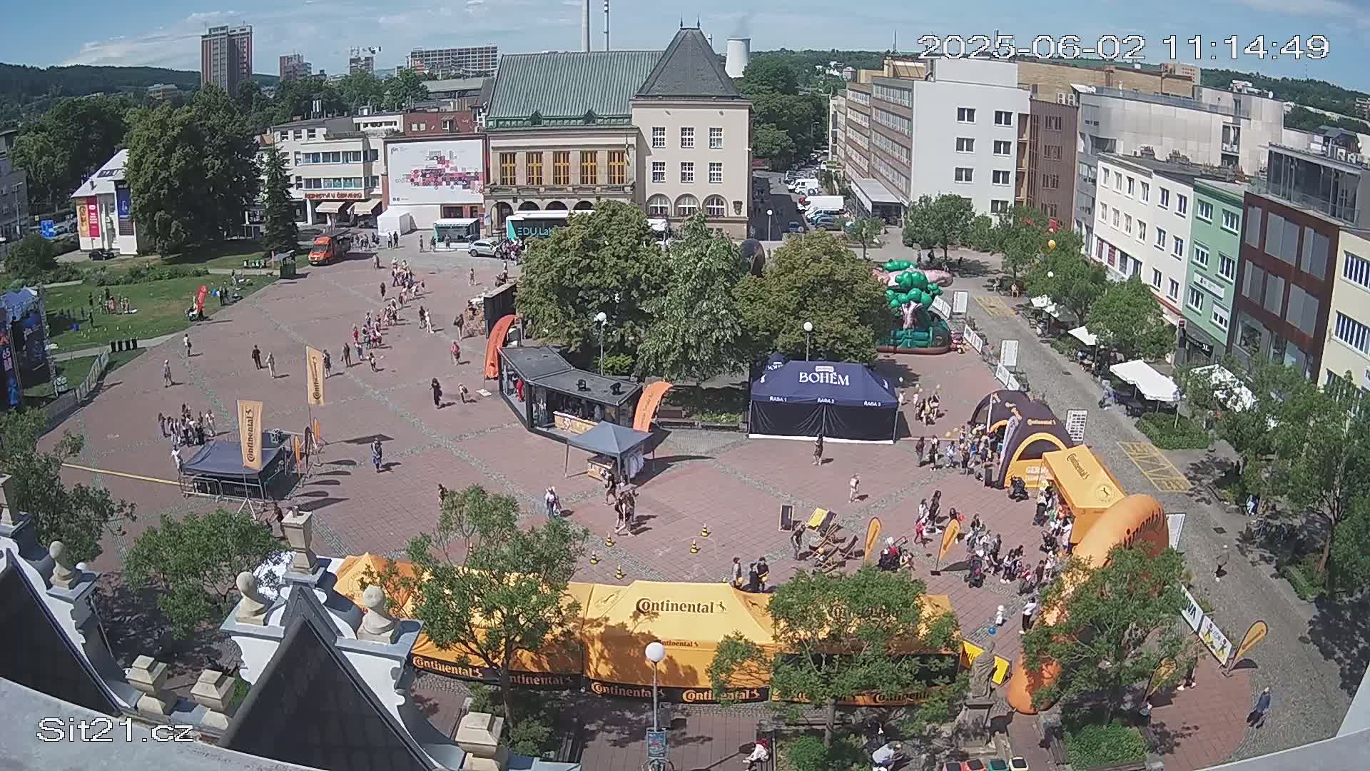A town square, filled with people and various vendor tents under a sunny sky, features several large inflatable structures.
