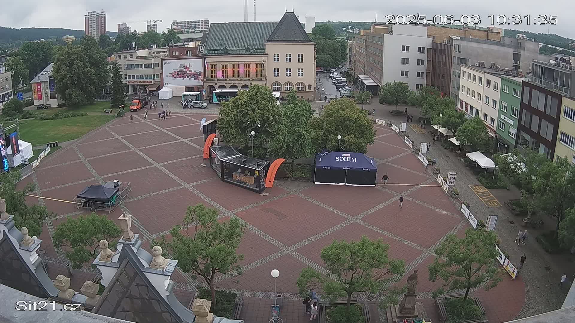 A brick town square with several tents, small crowds of people, and buildings surrounding it under an overcast sky.