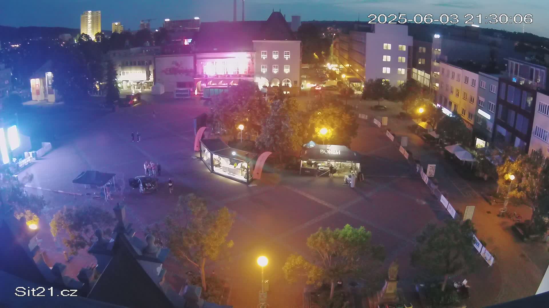 A nighttime, high-angle view of a town square with several small shops and kiosks, surrounded by buildings and trees under a dark sky.