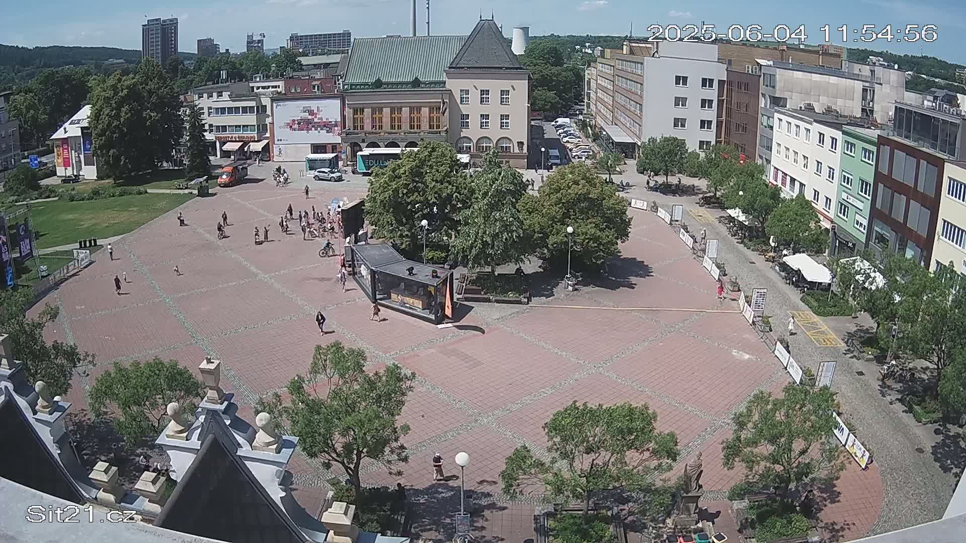 A sunny day shows a large town square with scattered people, buildings, and green spaces.