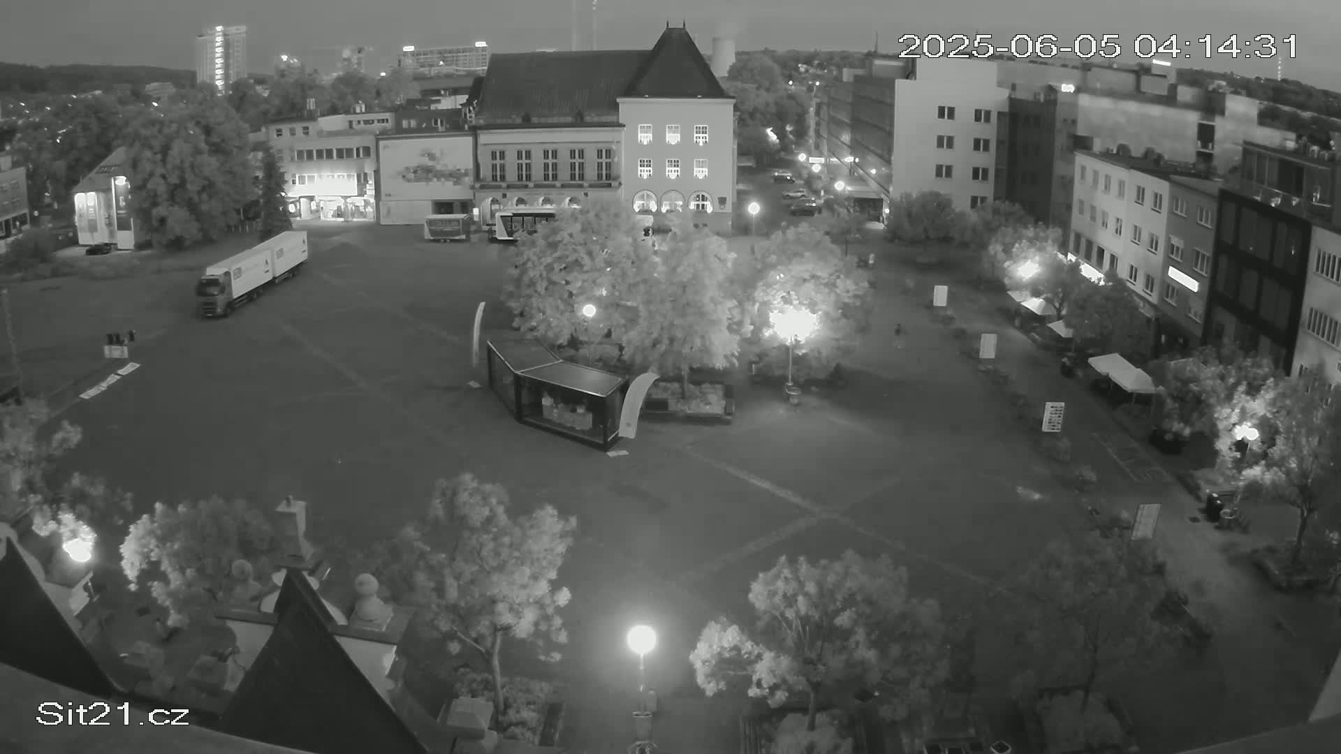 A nighttime, high-angle view of a town square with buildings surrounding it, trees in the center, and a truck parked nearby.