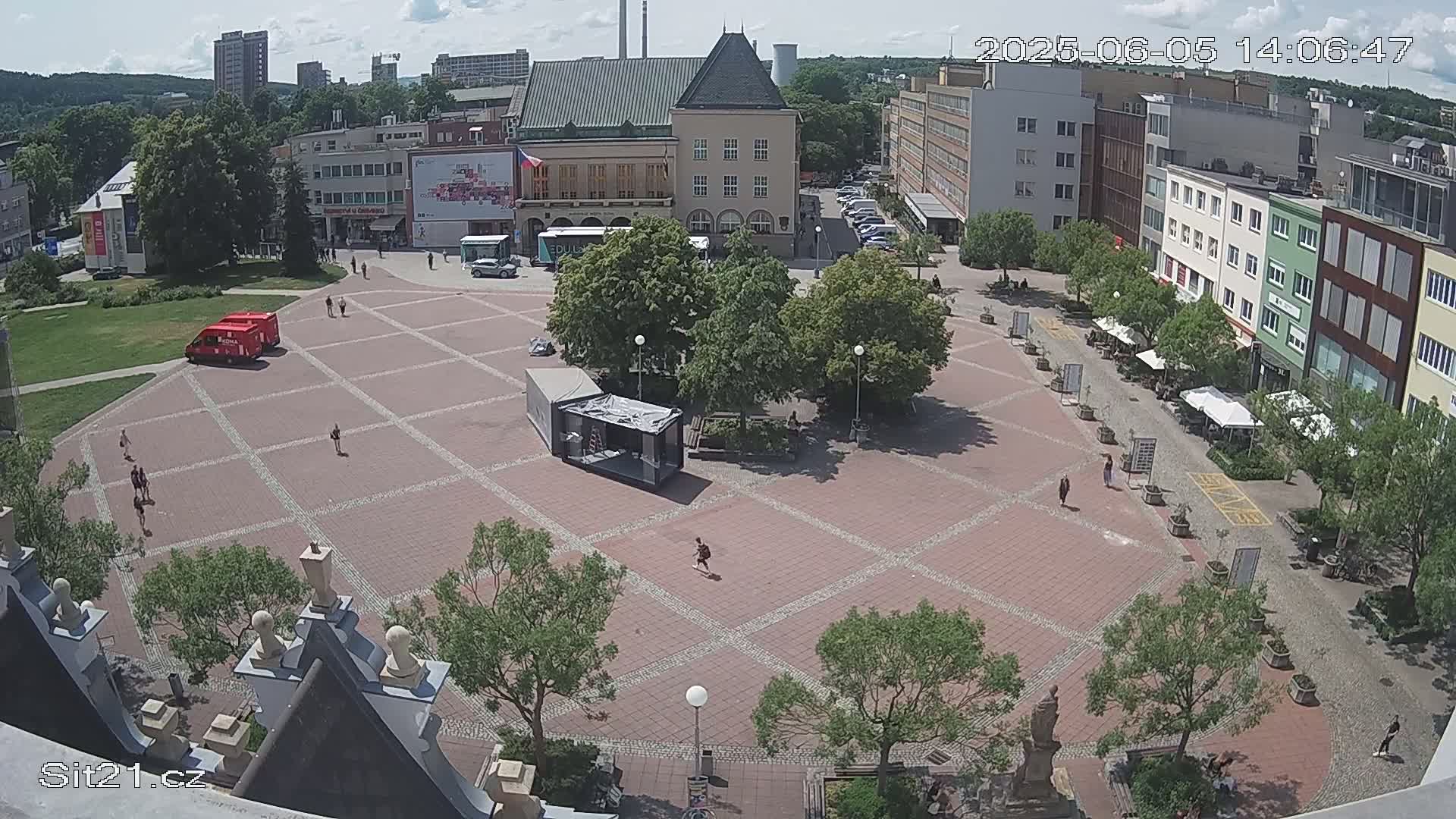 A mostly sunny, high-angle view of a large town square with several buildings surrounding it, some people walking around, and a few vehicles parked.