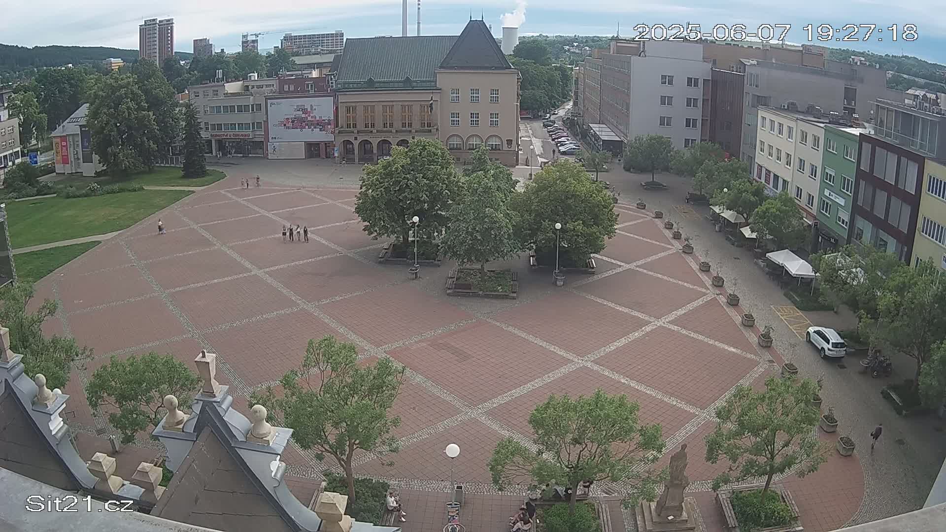 A large brick town square with several people and trees, under a partly cloudy sky.