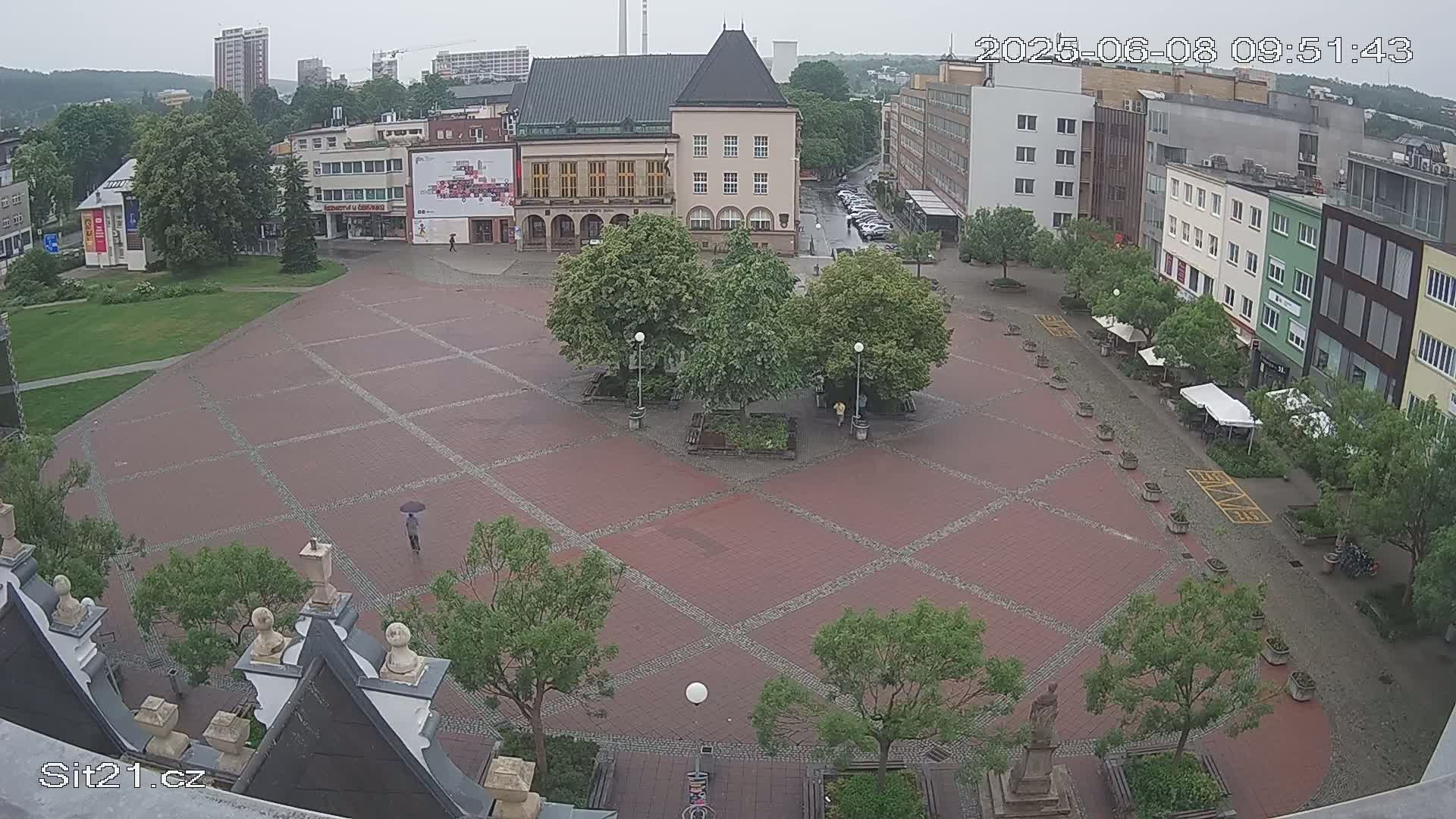 A brick town square is mostly empty under a rainy sky, with a few people and trees scattered around it, and buildings surrounding it.
