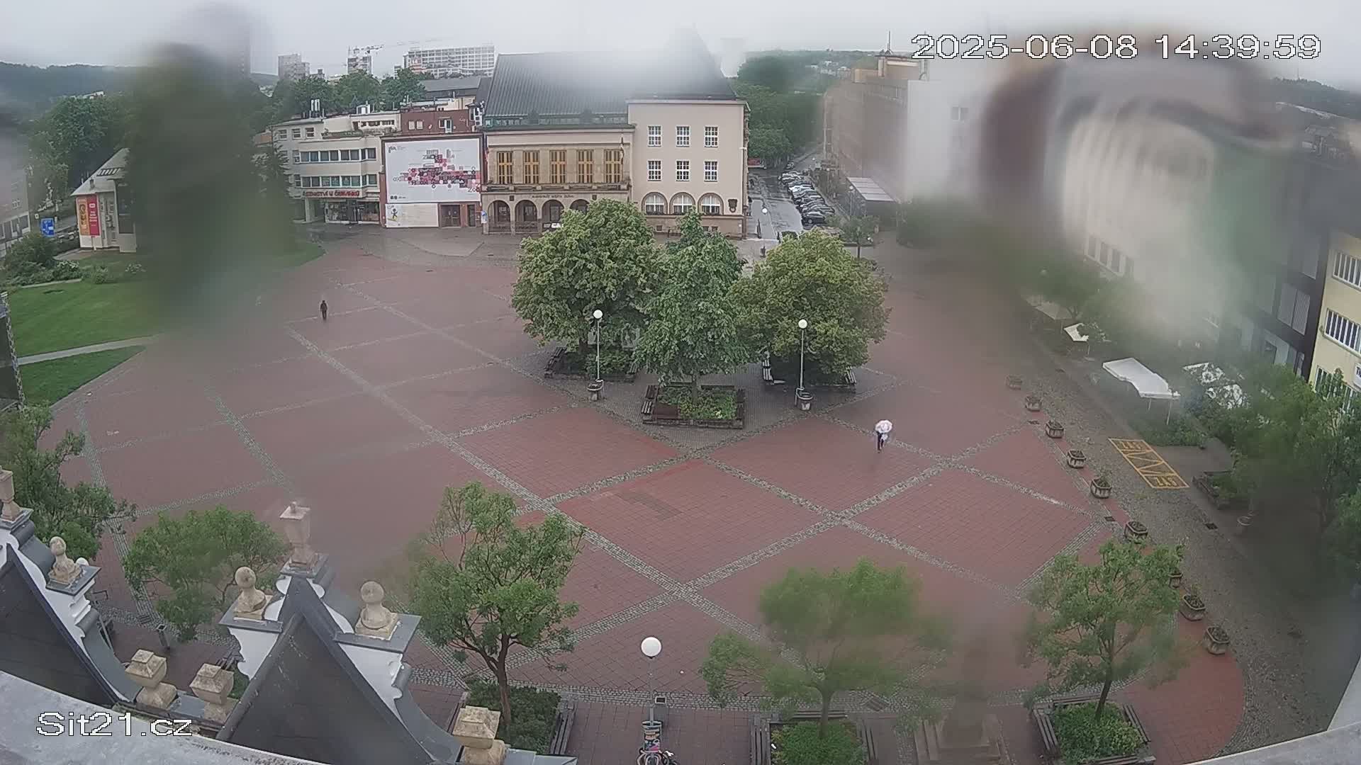 A rain-soaked brick town square is viewed from above, showing buildings, trees, and a few pedestrians with one using an umbrella.