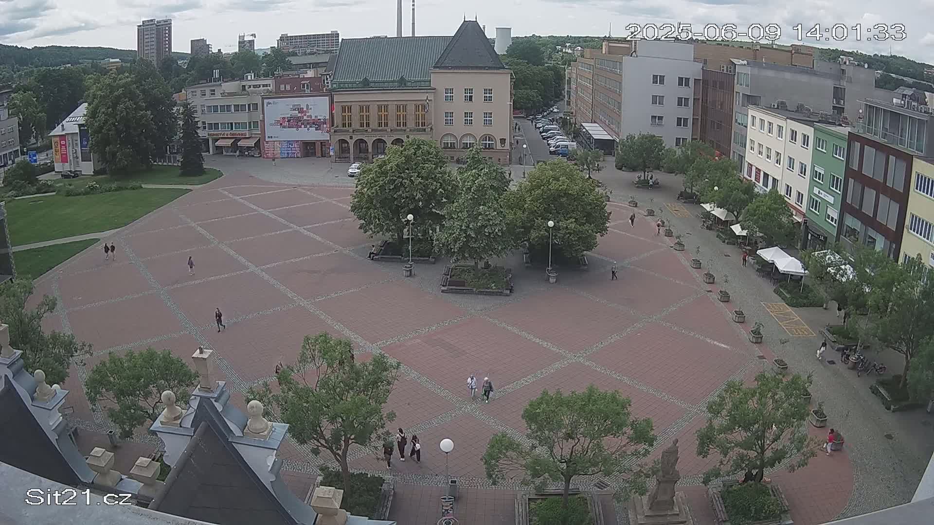 A brick town square with several people walking around, surrounded by buildings and trees under a partly cloudy sky.