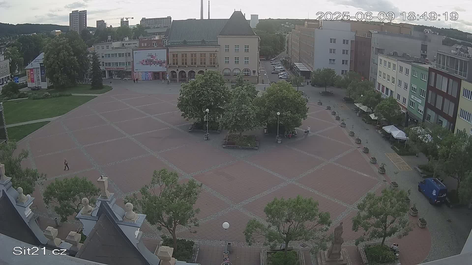 An overcast day reveals a large, paved town square with several trees and buildings surrounding it, a few people are visible.