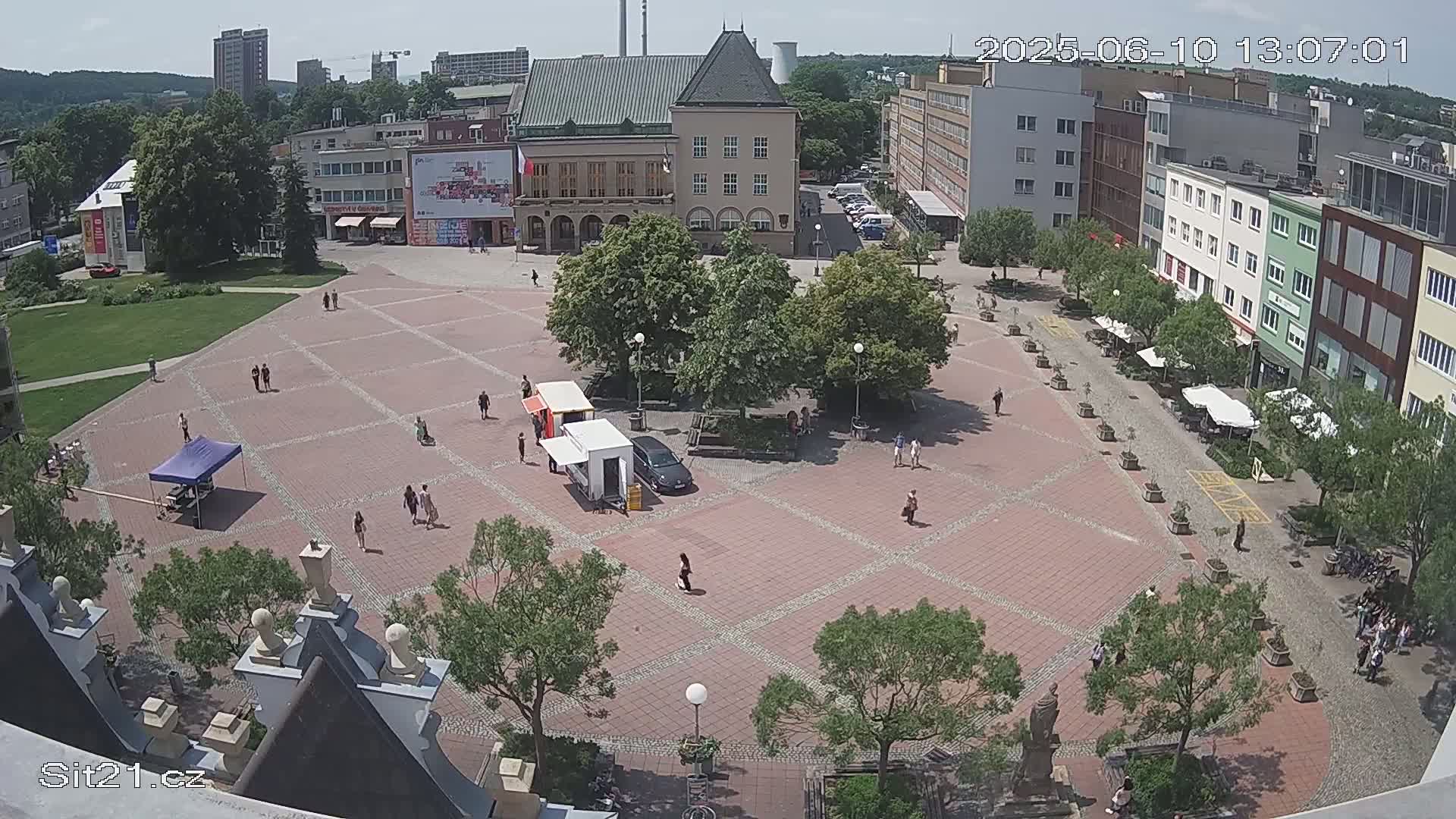A brick town square with several people and small vendors is surrounded by buildings and trees on a sunny day.