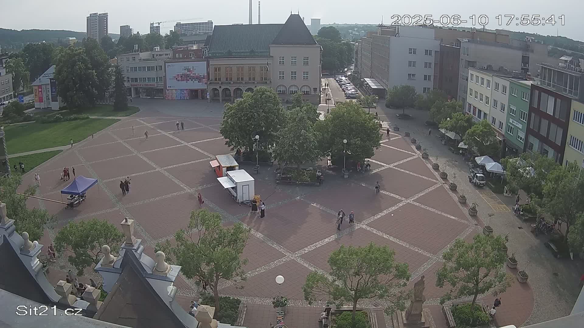 A mostly sunny overhead view of a town square with several people scattered throughout, surrounded by buildings and trees.