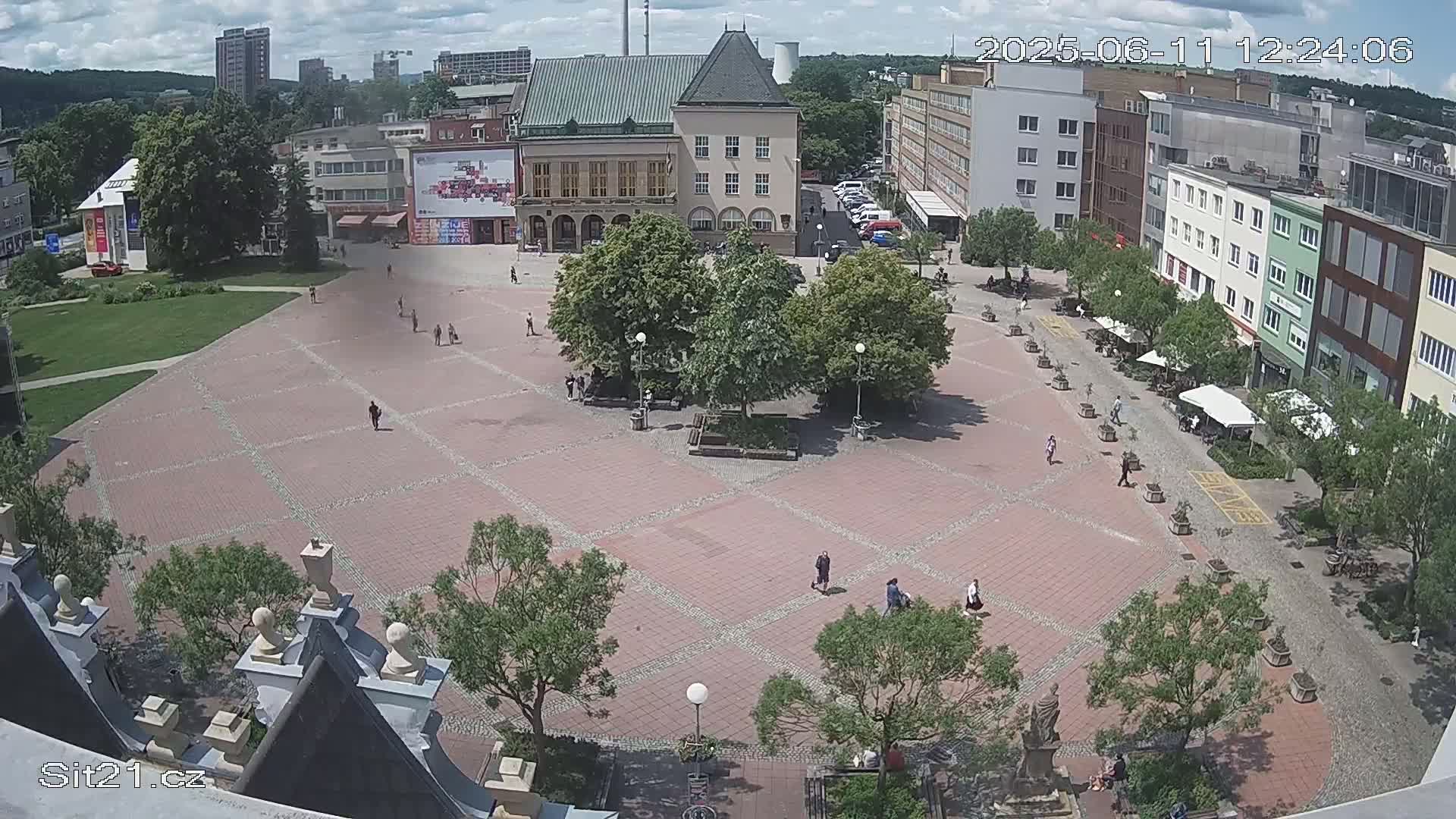 A large, brick-paved town square with several buildings surrounding it, some trees and people scattered throughout, on a partly sunny day.