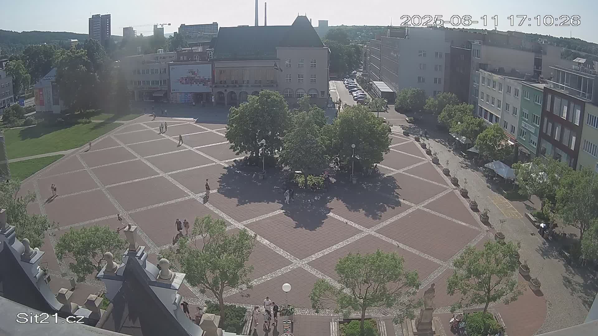A sunny, high-angle view shows a large, brick plaza with scattered people, trees, and surrounding buildings.