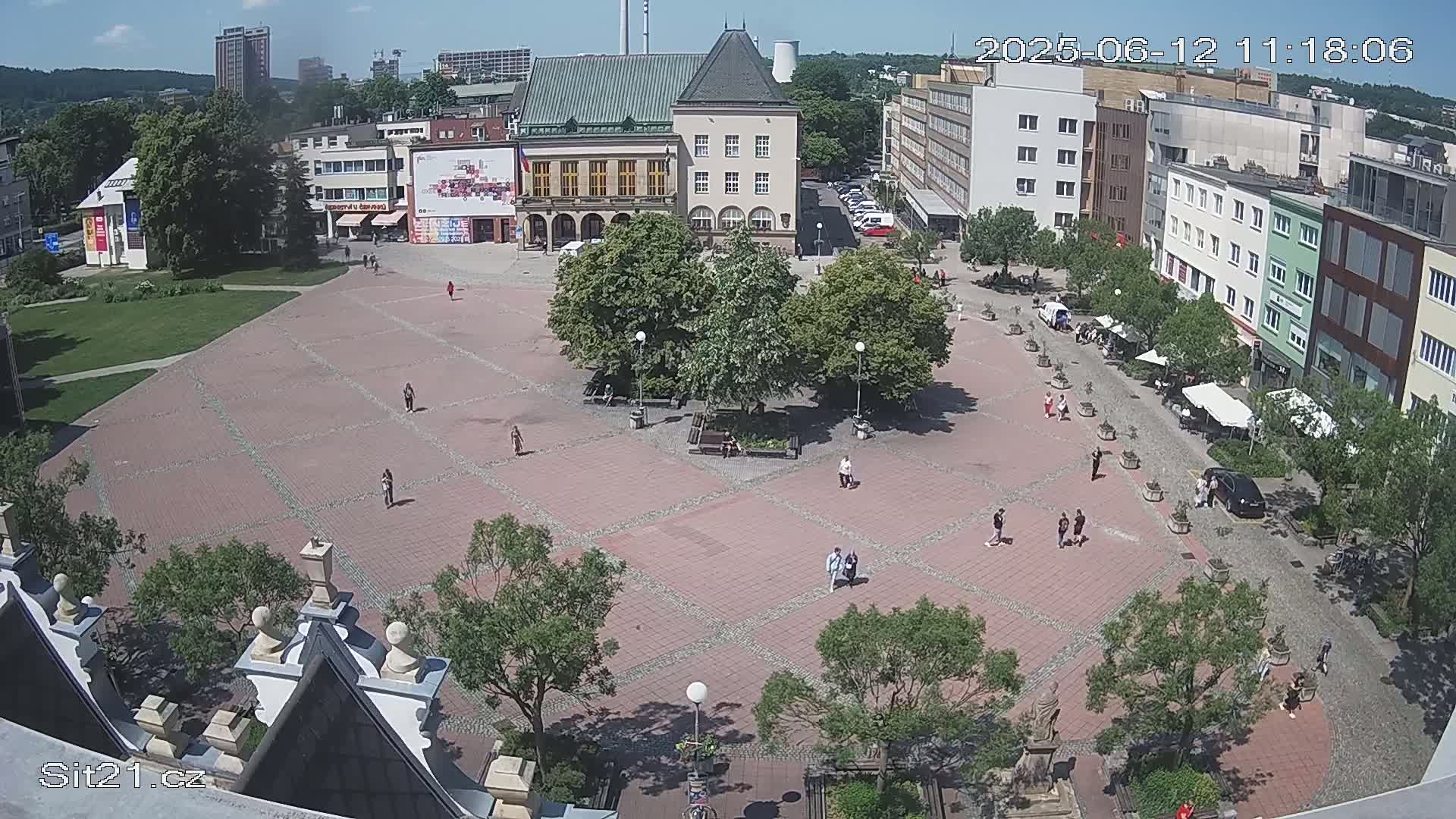 A high-angle view of a town square on a sunny day shows people walking on a brick plaza surrounded by buildings and trees.