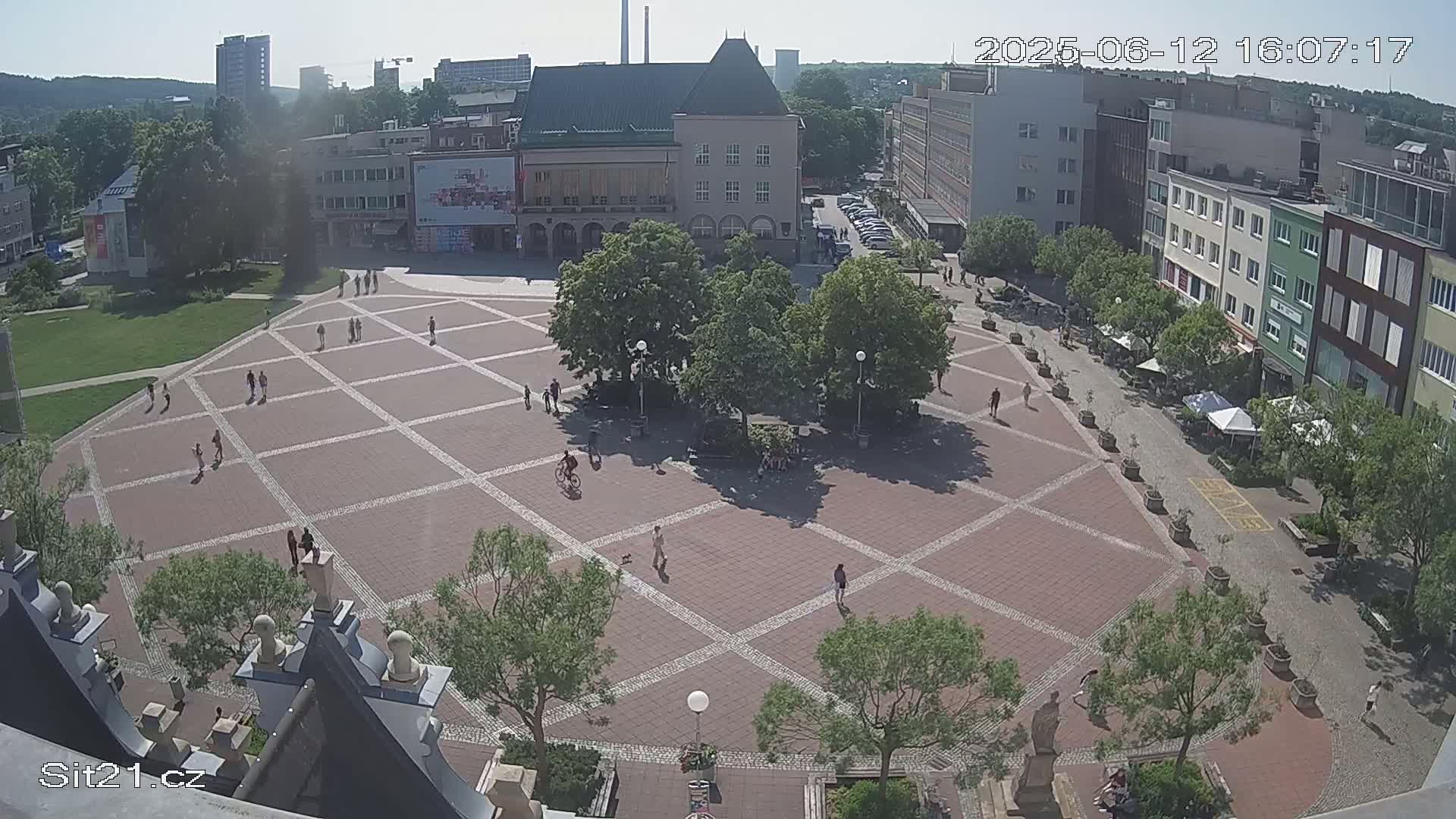 A large, brick plaza with scattered pedestrians and trees is surrounded by buildings on a sunny day.