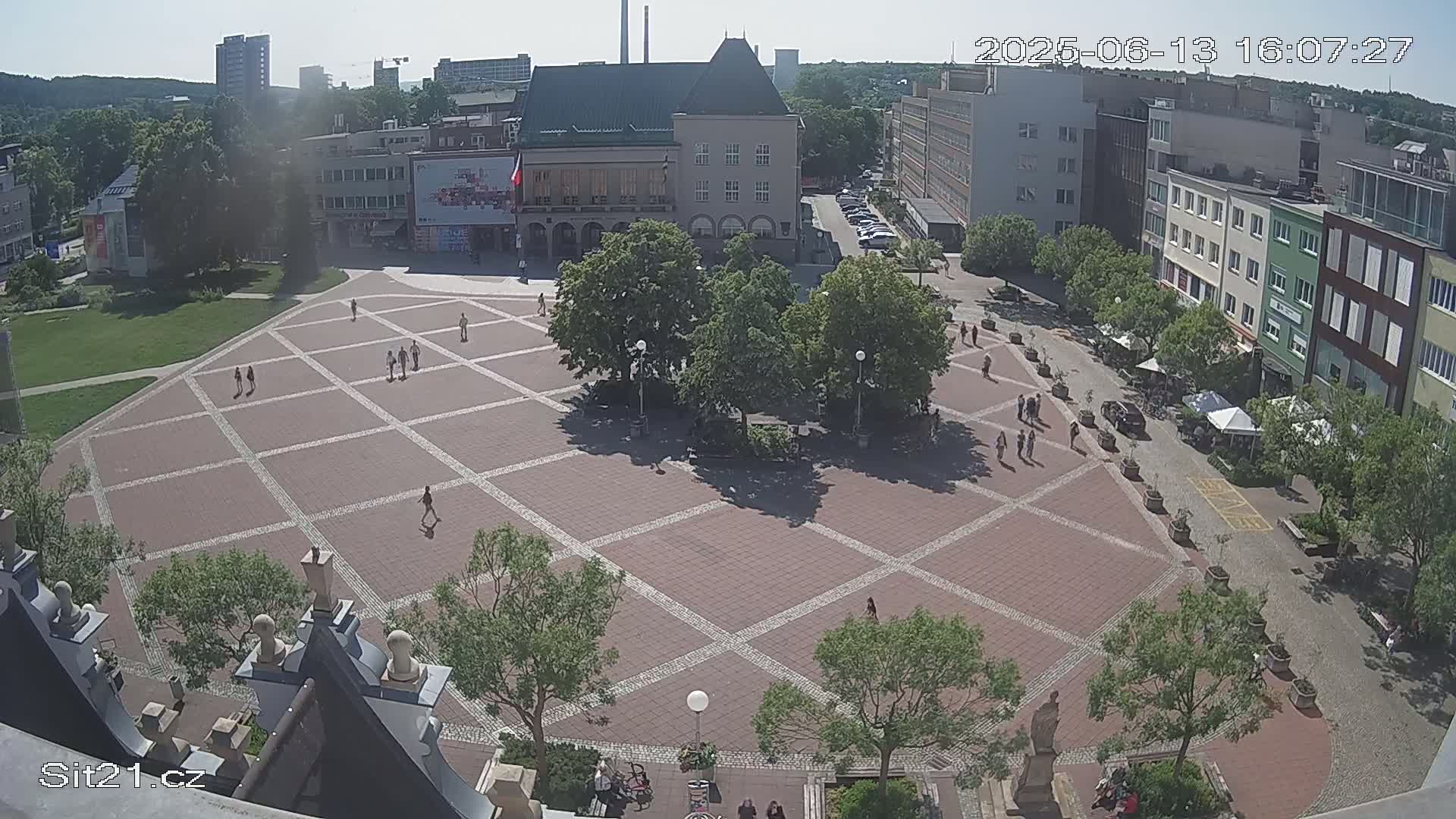 A large, paved town square with a grid pattern, surrounded by buildings and trees, on a sunny day.