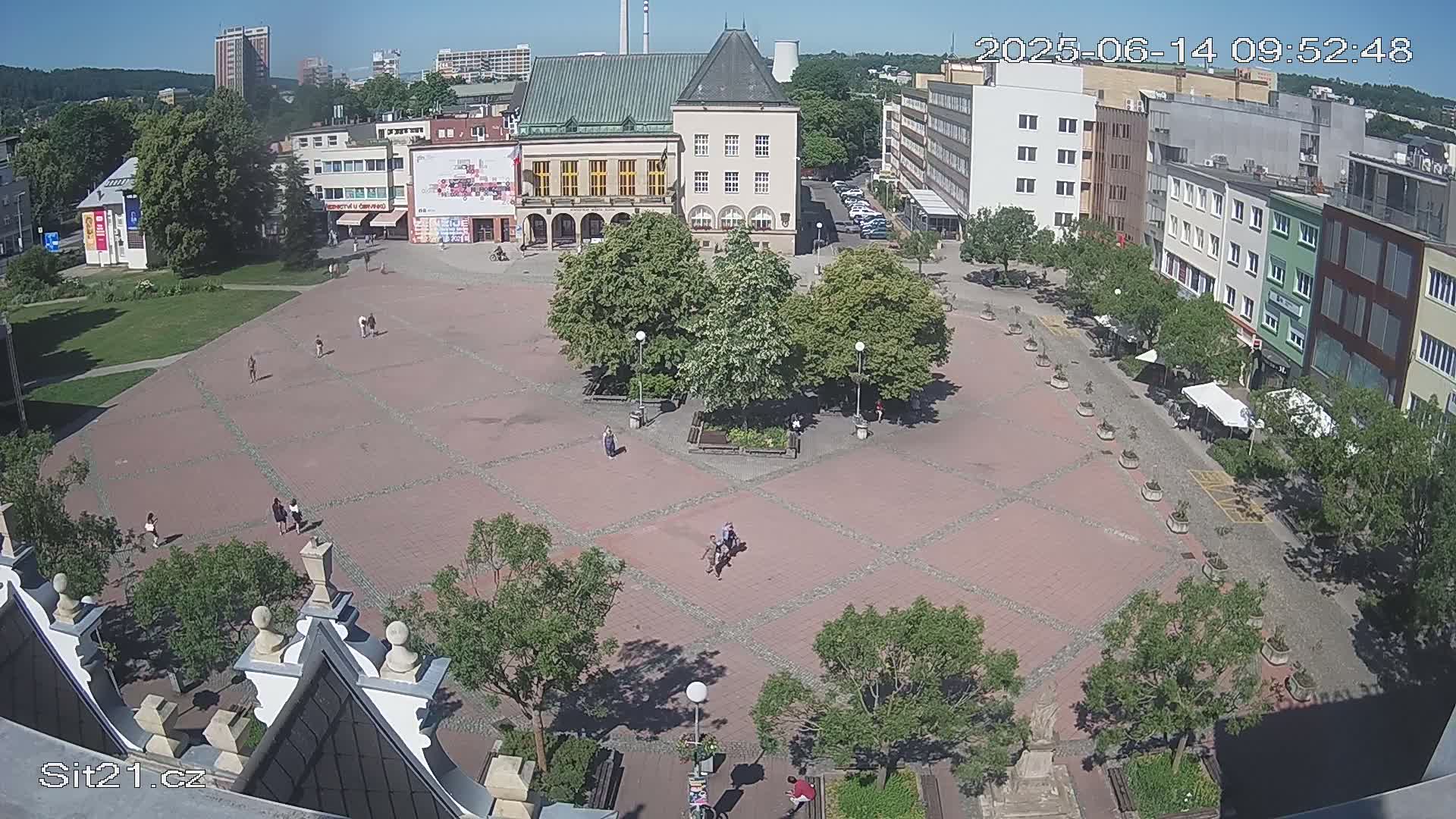 A large town square with a brick paving pattern, several trees, and a few people is seen from above on a sunny day.