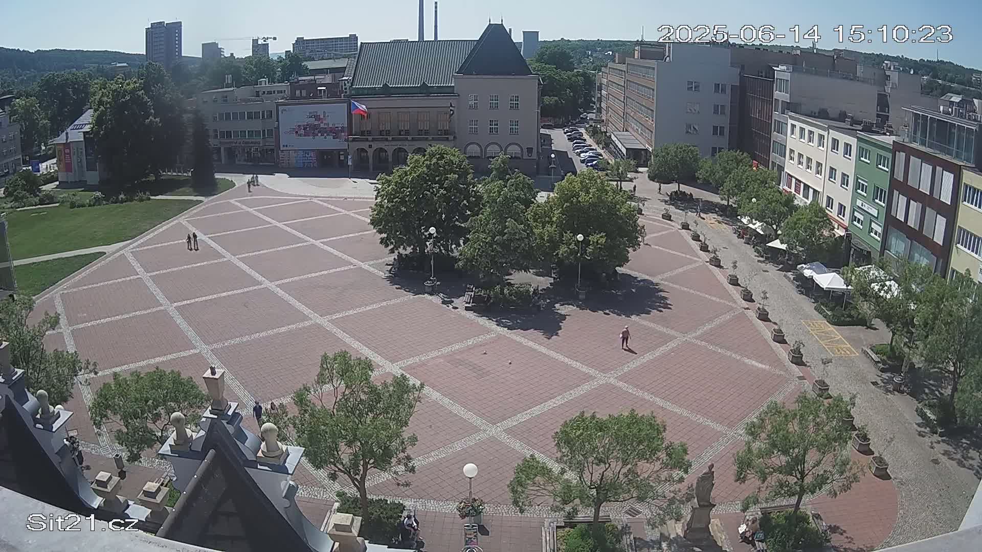 A large, brick-paved town square with several buildings and trees surrounding it on a sunny day.