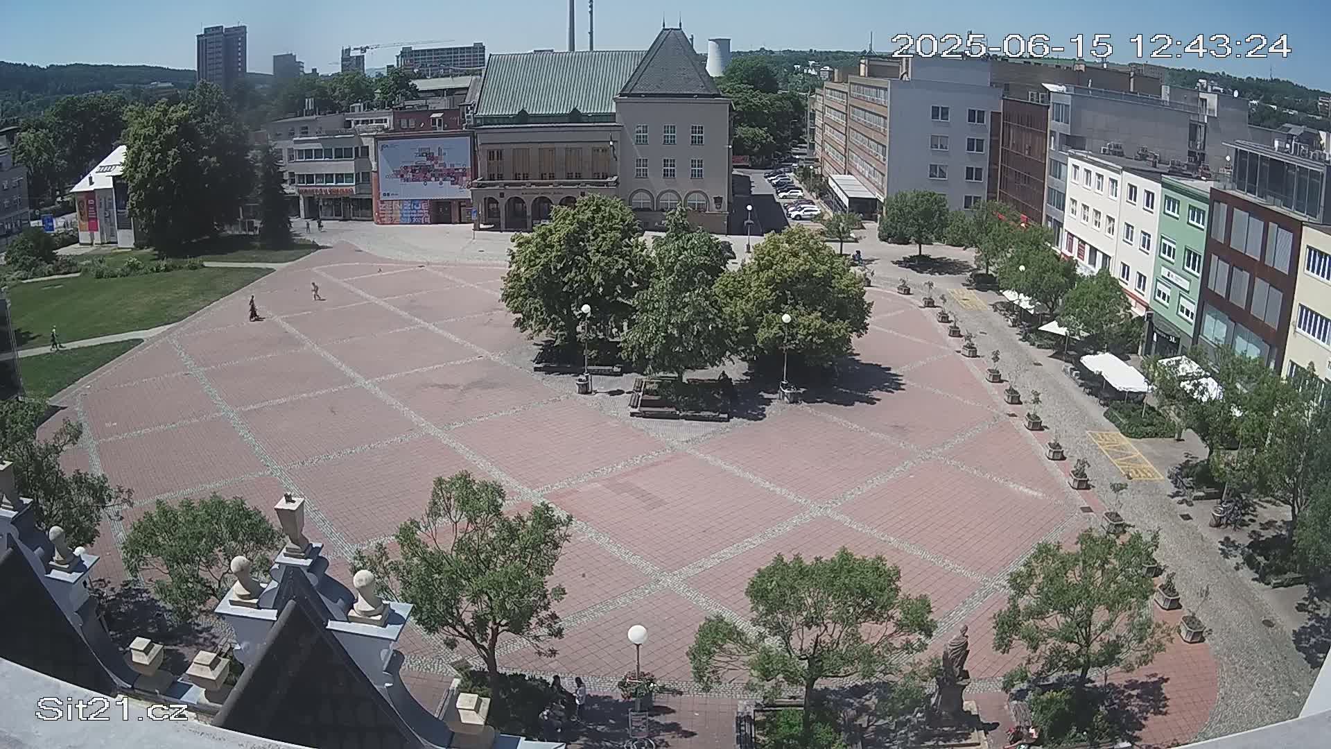 An aerial view of a brick town square on a sunny day, surrounded by buildings and trees, with a few people visible.