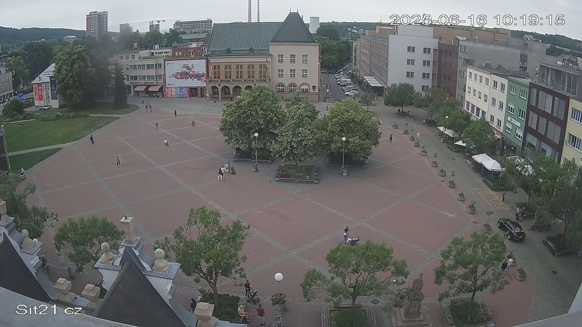 An overcast day shows a large, brick town square with several people walking around and sitting near trees and buildings.