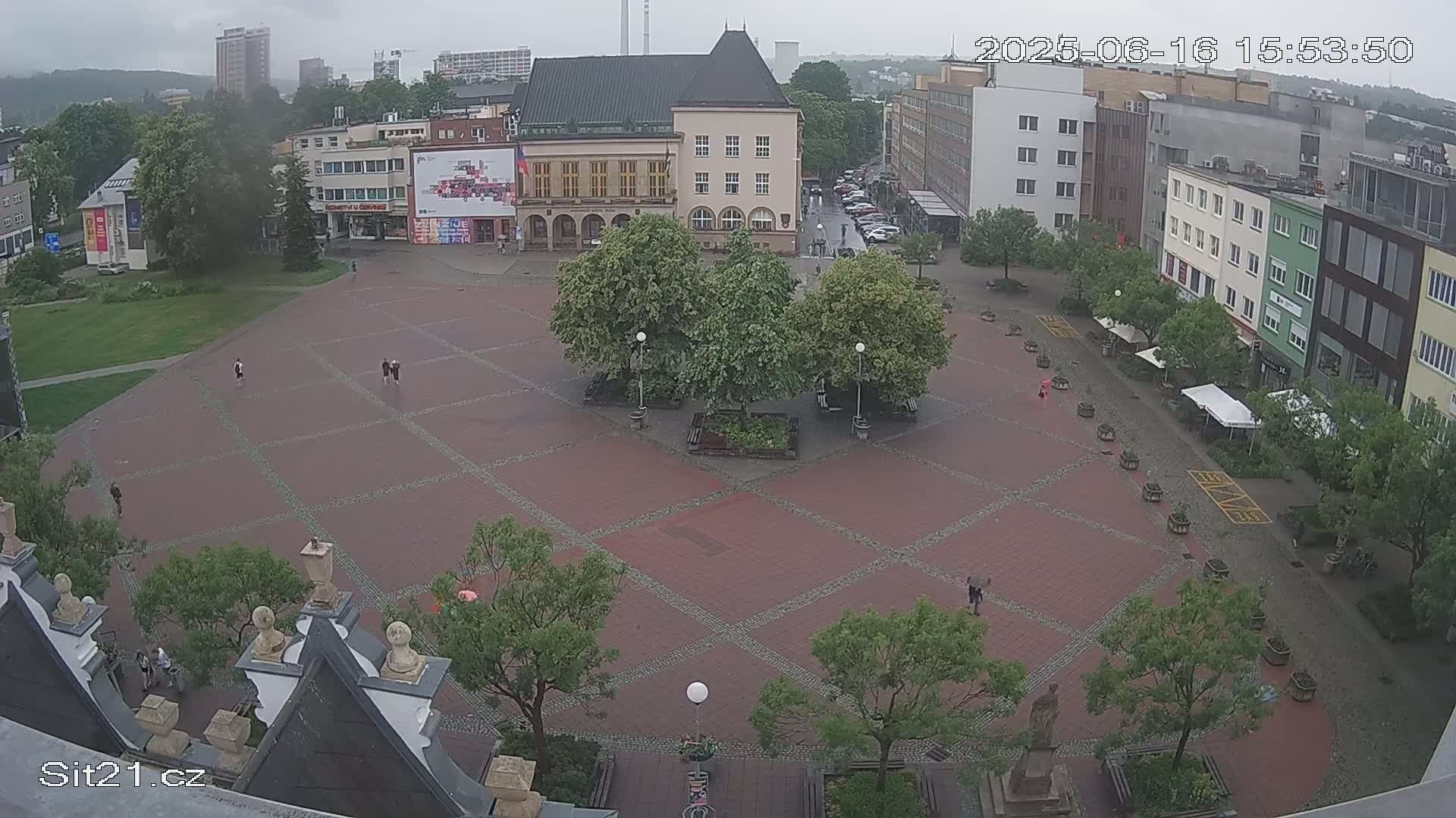 An overcast day reveals a brick town square with several people walking and trees planted throughout, surrounded by buildings.