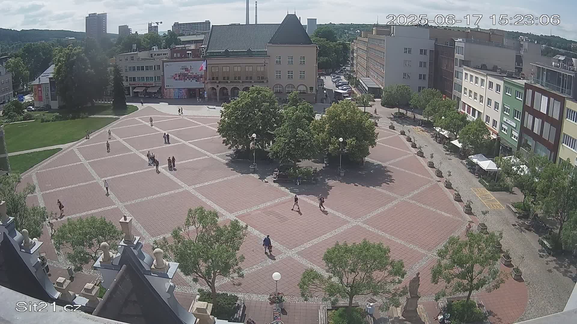 A large, brick town square with a few people walking around, surrounded by buildings and trees on a sunny day.