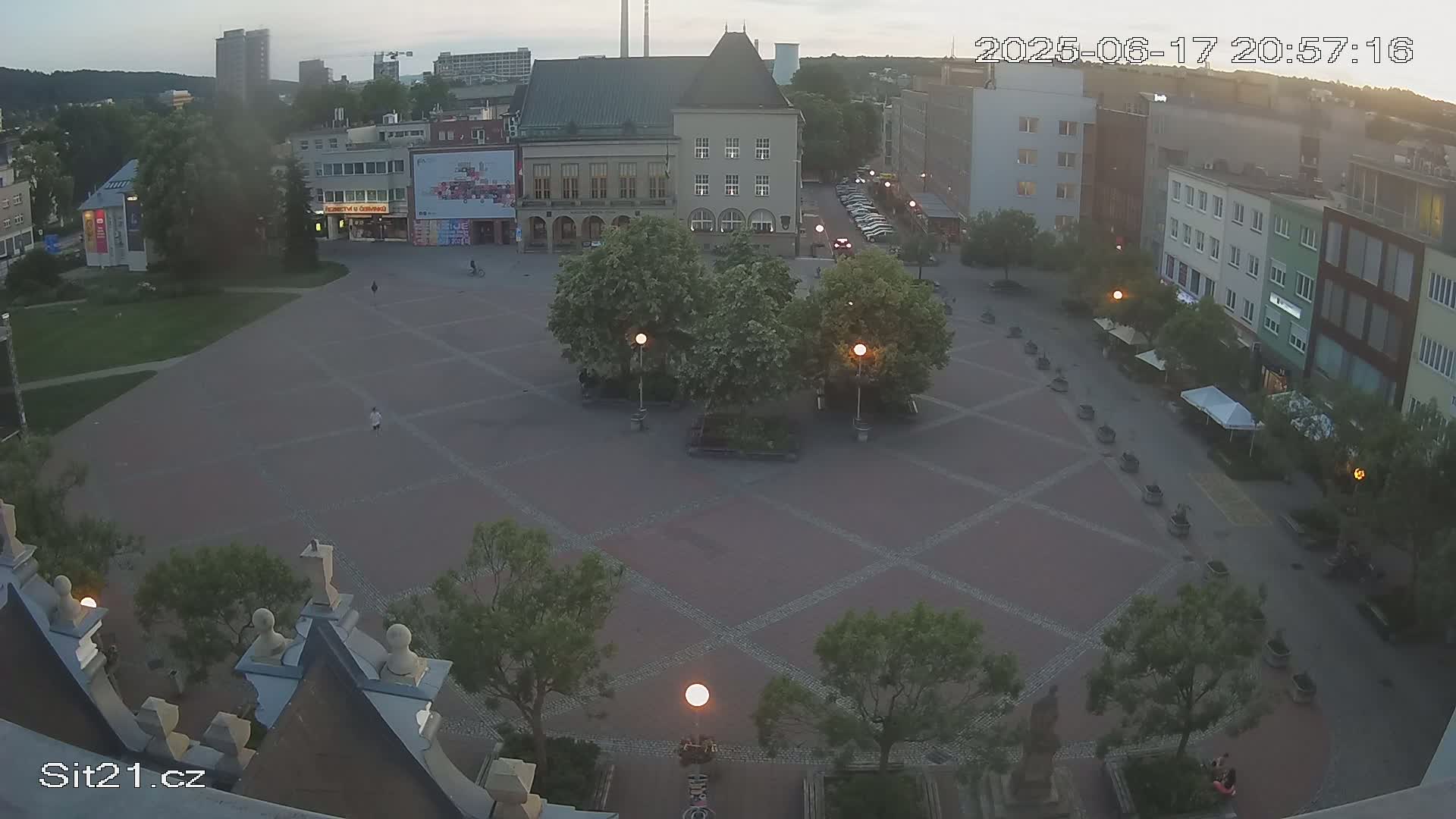 An empty town square at dusk, under a mostly clear sky, surrounded by buildings and trees.
