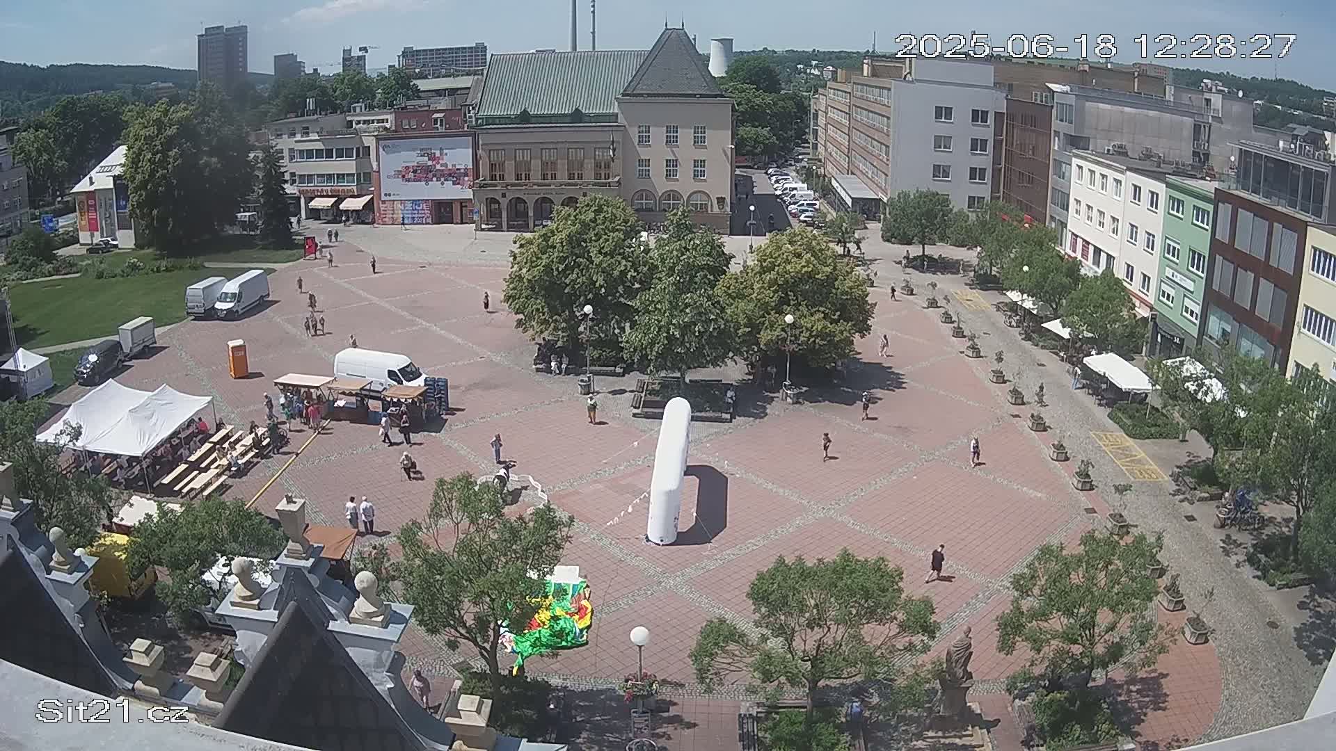 A sunny day shows a town square with people, trees, buildings, and market stalls.