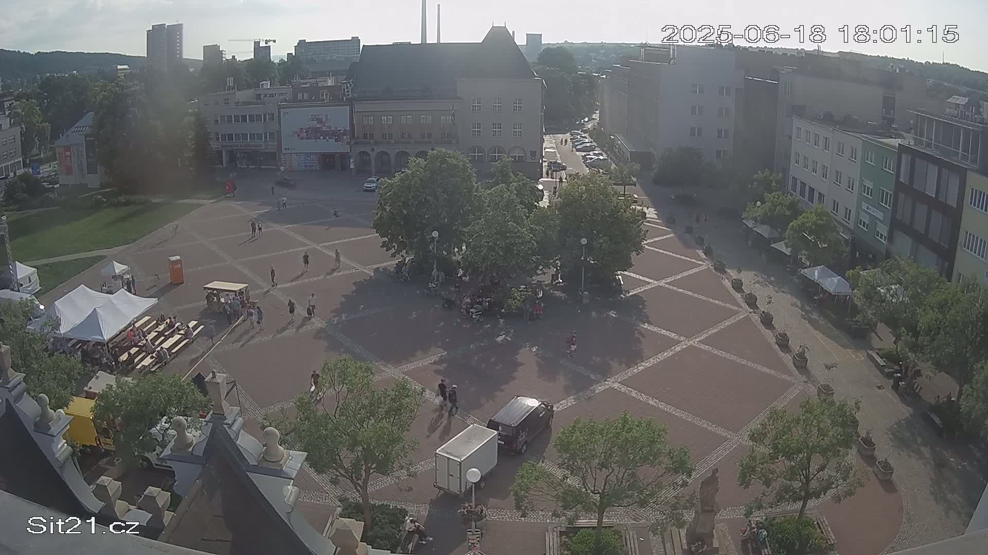 A paved town square with scattered people, trees, and small tents under a partly sunny sky.