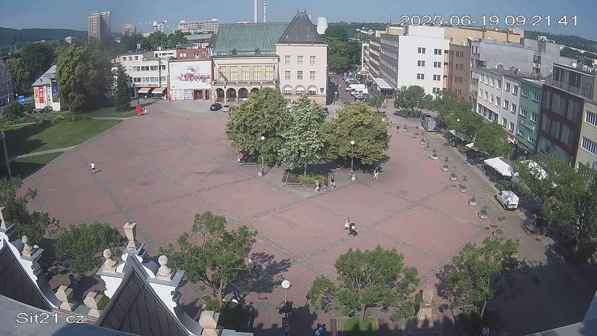 A brick town square with several buildings surrounding it and trees providing shade on a sunny day.