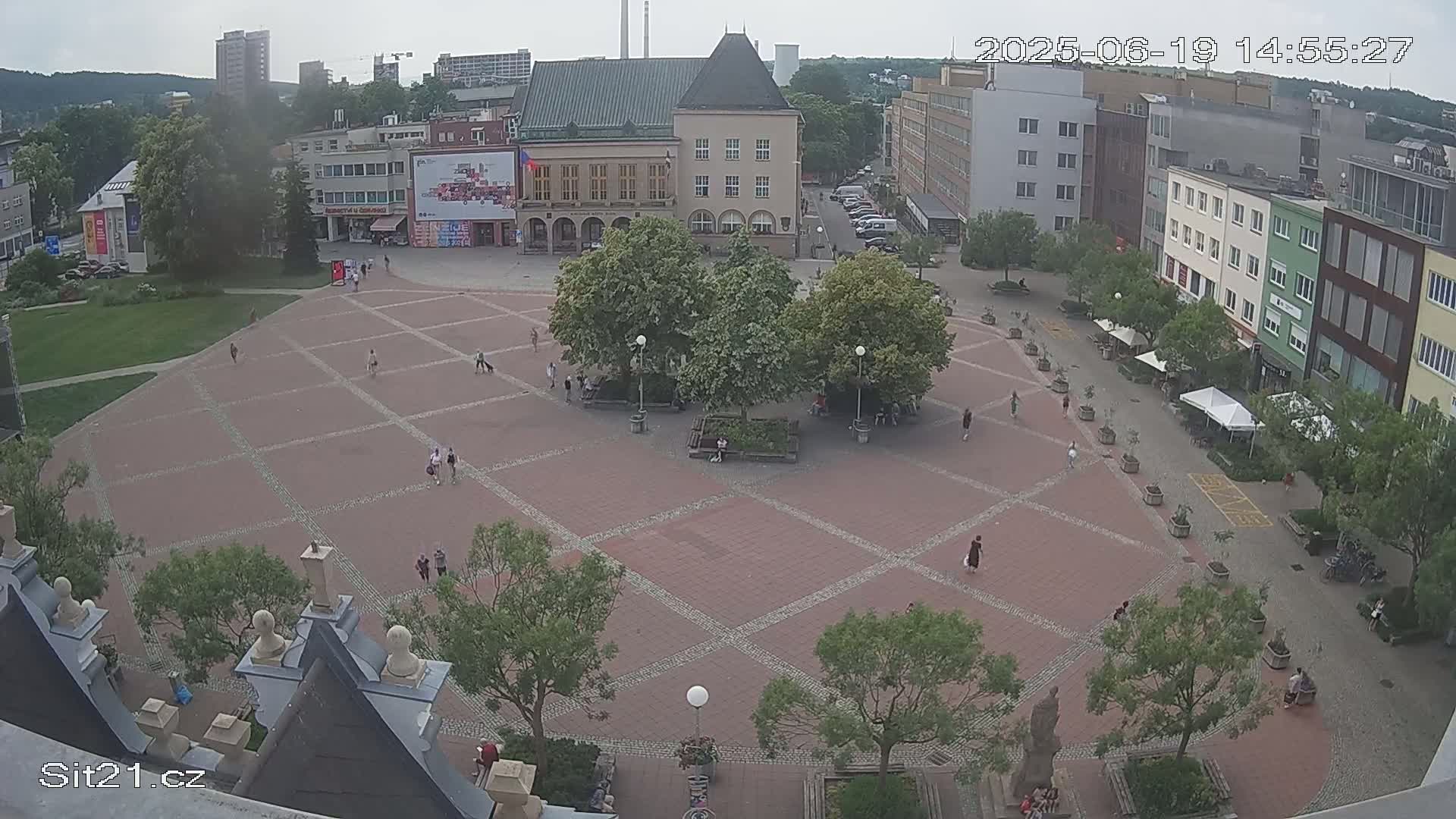 A brick town square with sparsely scattered people, surrounded by buildings and trees under an overcast sky.