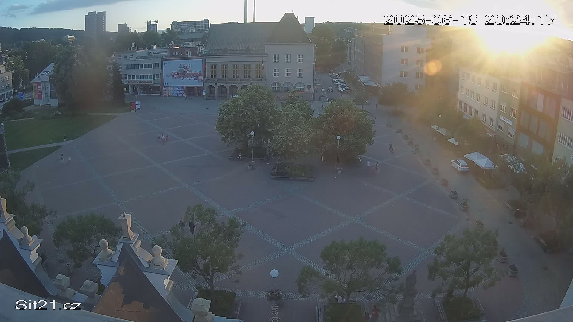 An aerial view of a town square at sunset, showing several buildings and trees, with a few people scattered throughout.