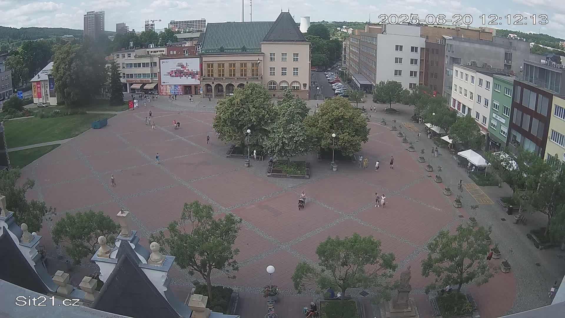 A brick town square with scattered pedestrians and trees under a partly cloudy sky.