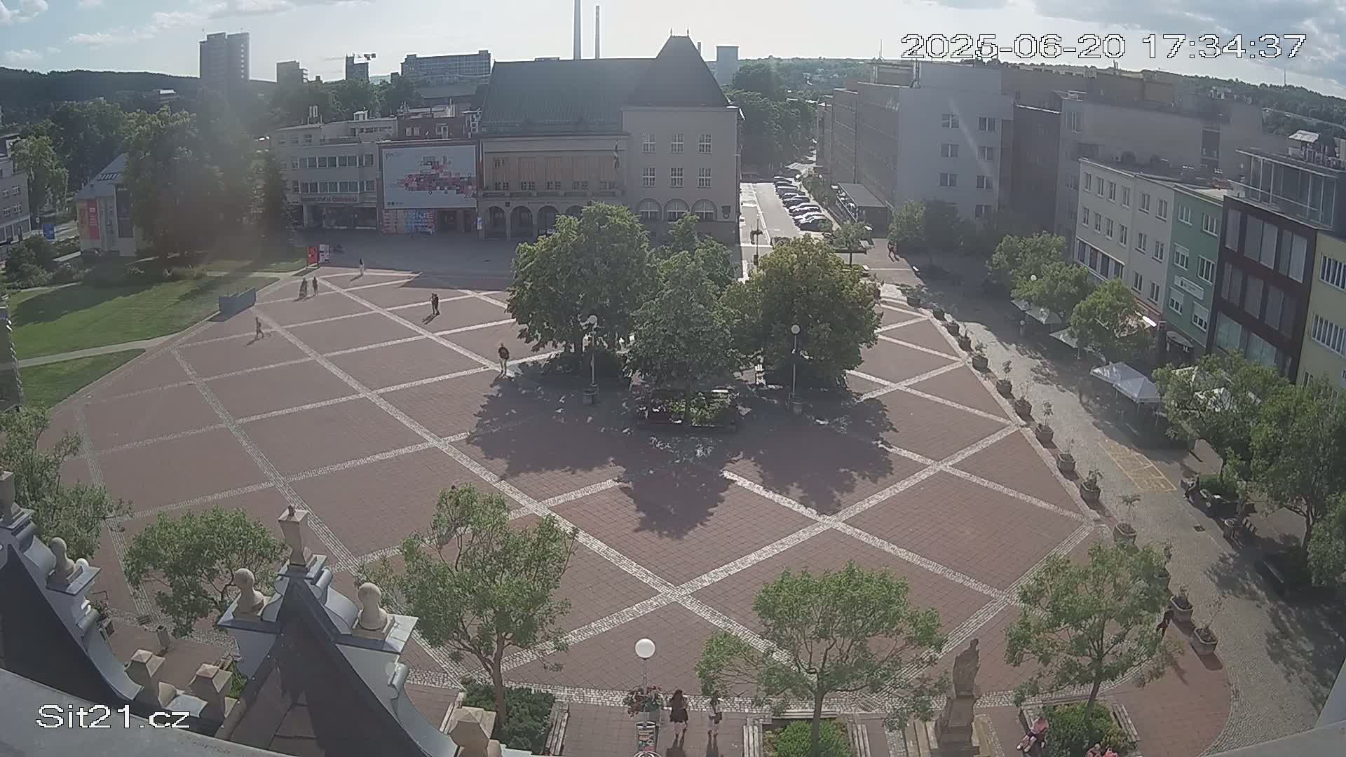 A paved square with several trees and a few people is seen from above on a partly sunny day.