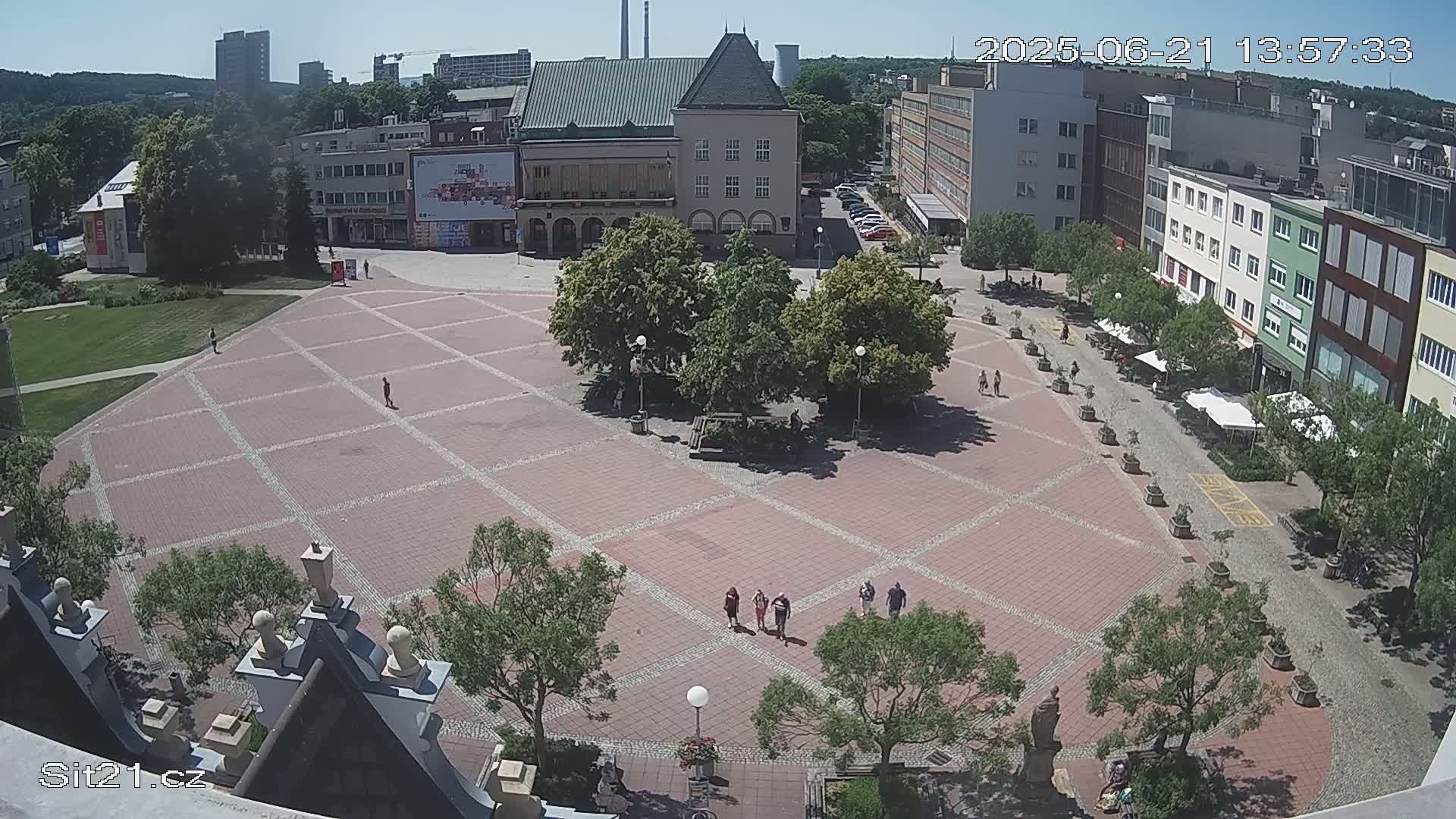 A large, brick plaza with several trees and a few people walking, surrounded by buildings under a sunny sky.